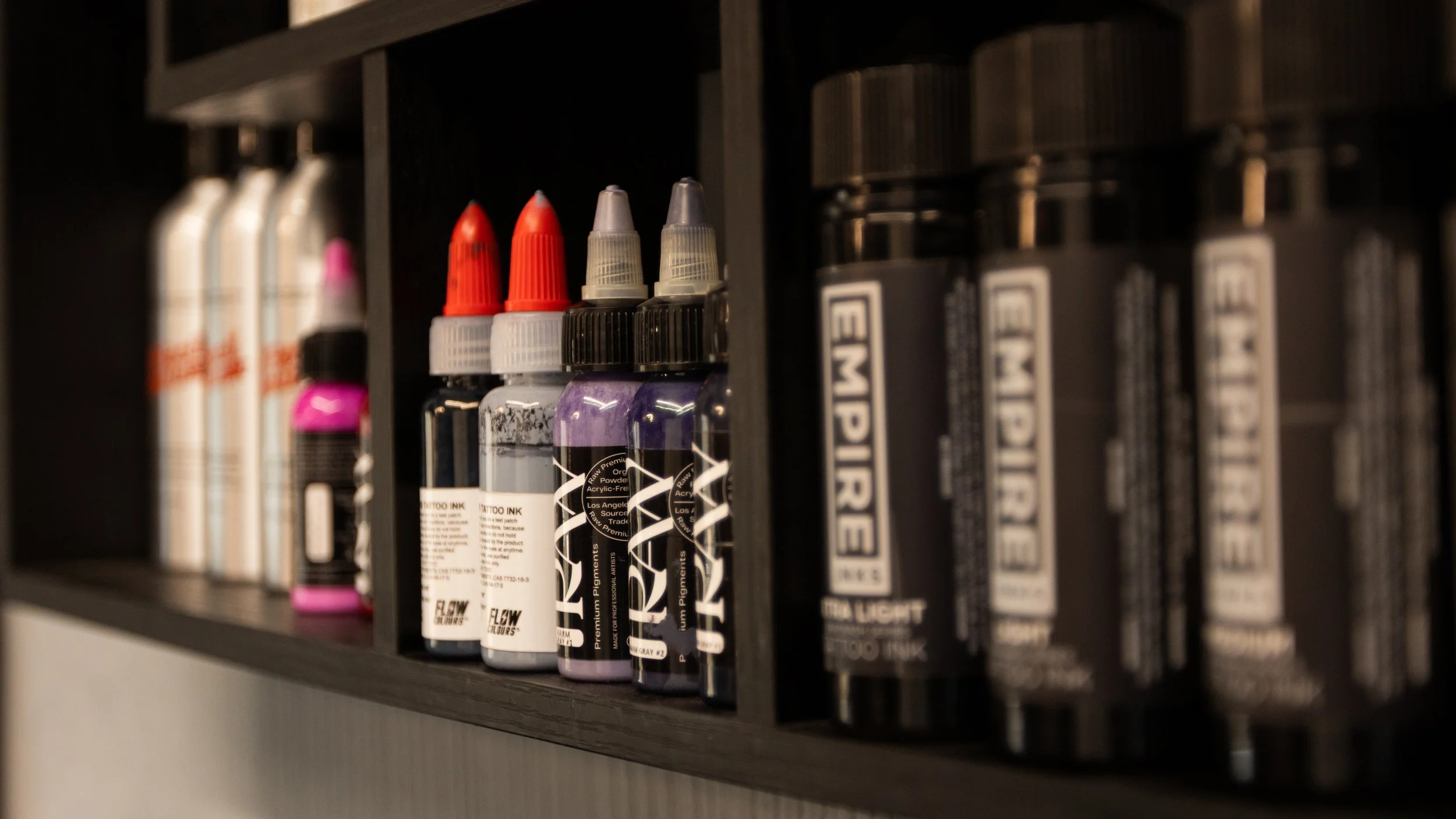 Shelf displaying bottles of tattoo ink in various colors, with other tattoo supply bottles on either side.