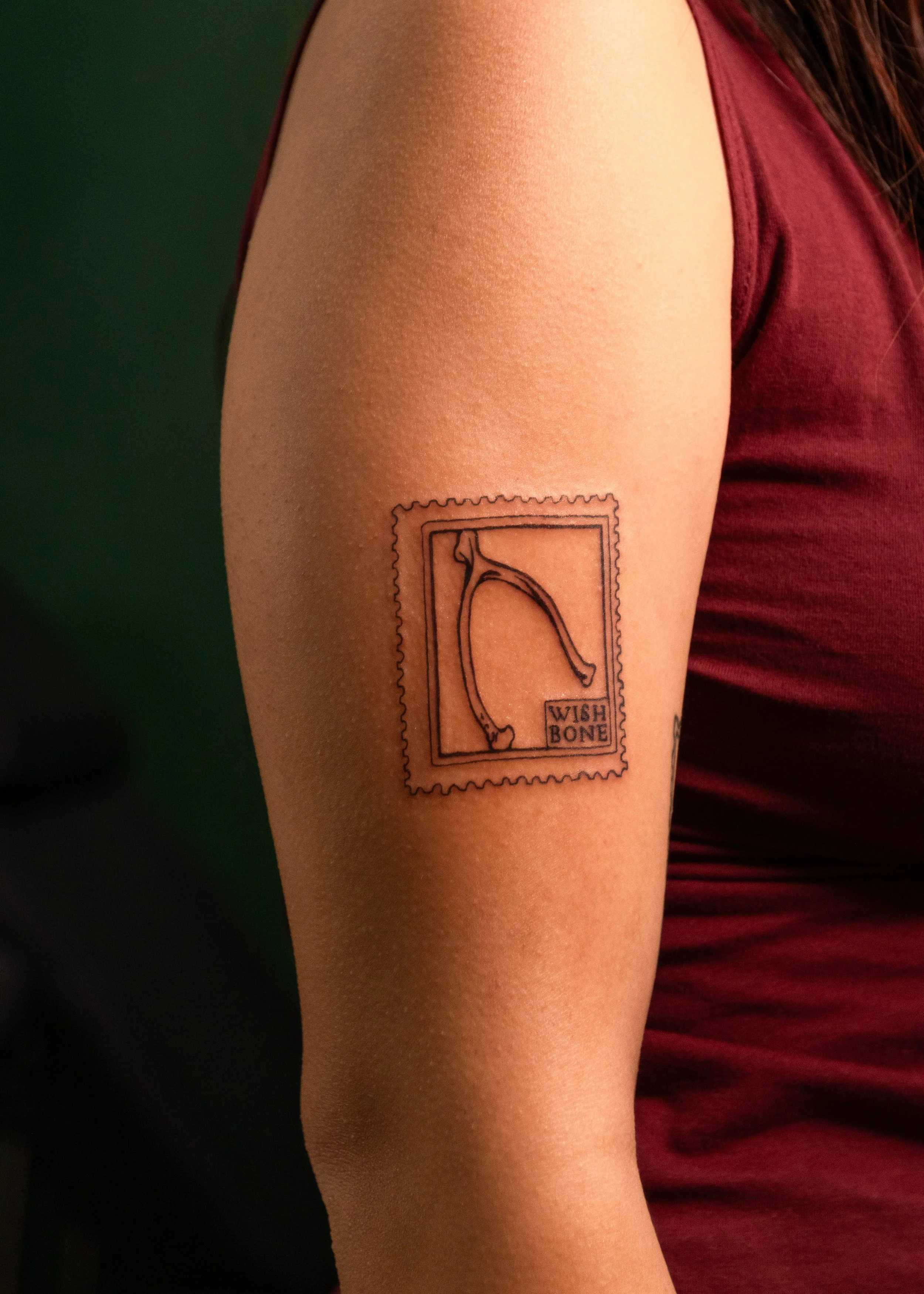 A person with fair skin showing a tattoo of a wishbone on their upper arm. The tattoo is styled as a postage stamp with the words "WISH BONE" written in a box below the wishbone.