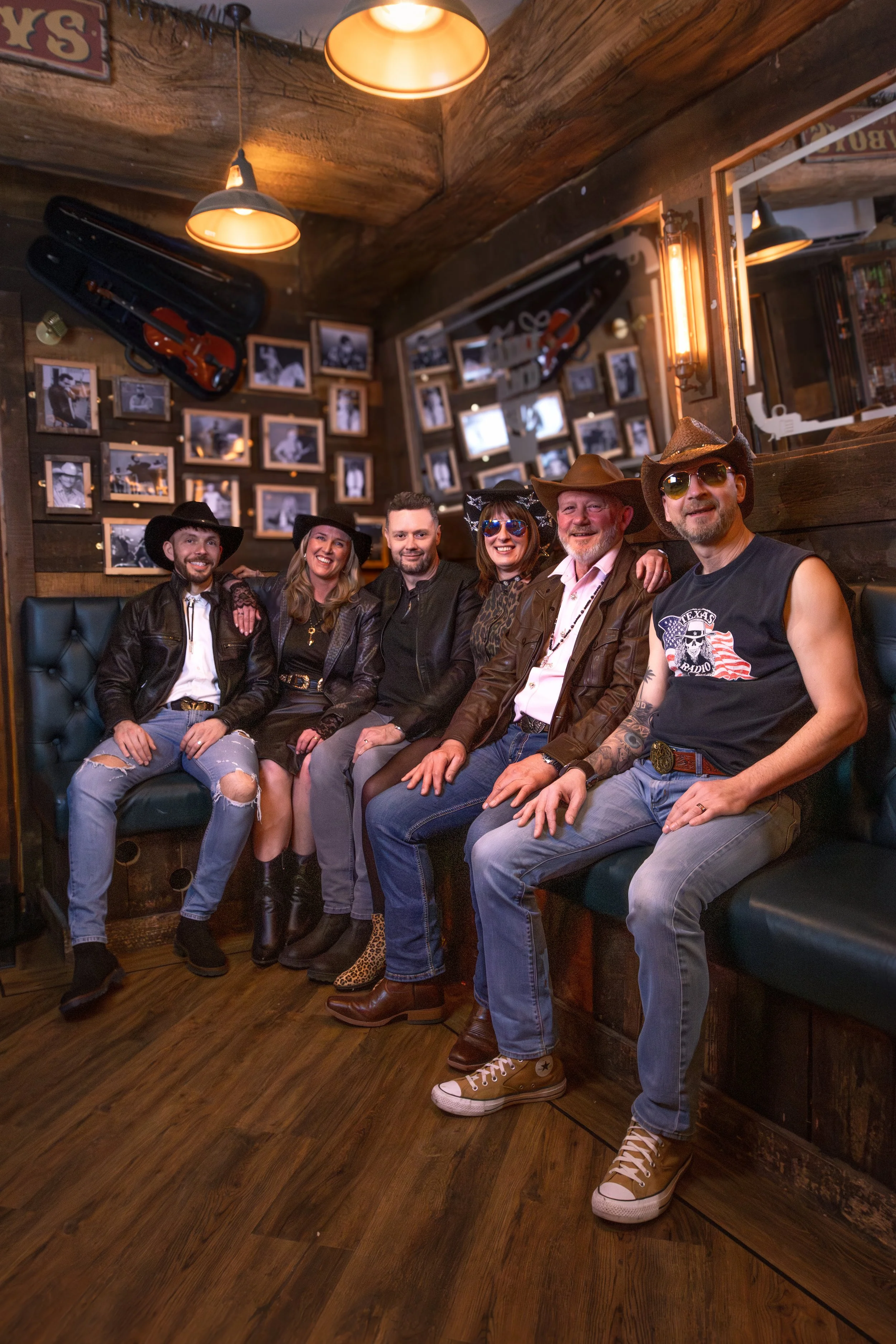Six people sitting on a booth in a bar, smiling and wearing cowboy hats. The background features a wall with black and white photographs and a guitar case hanging on it. The lighting is warm and rustic.