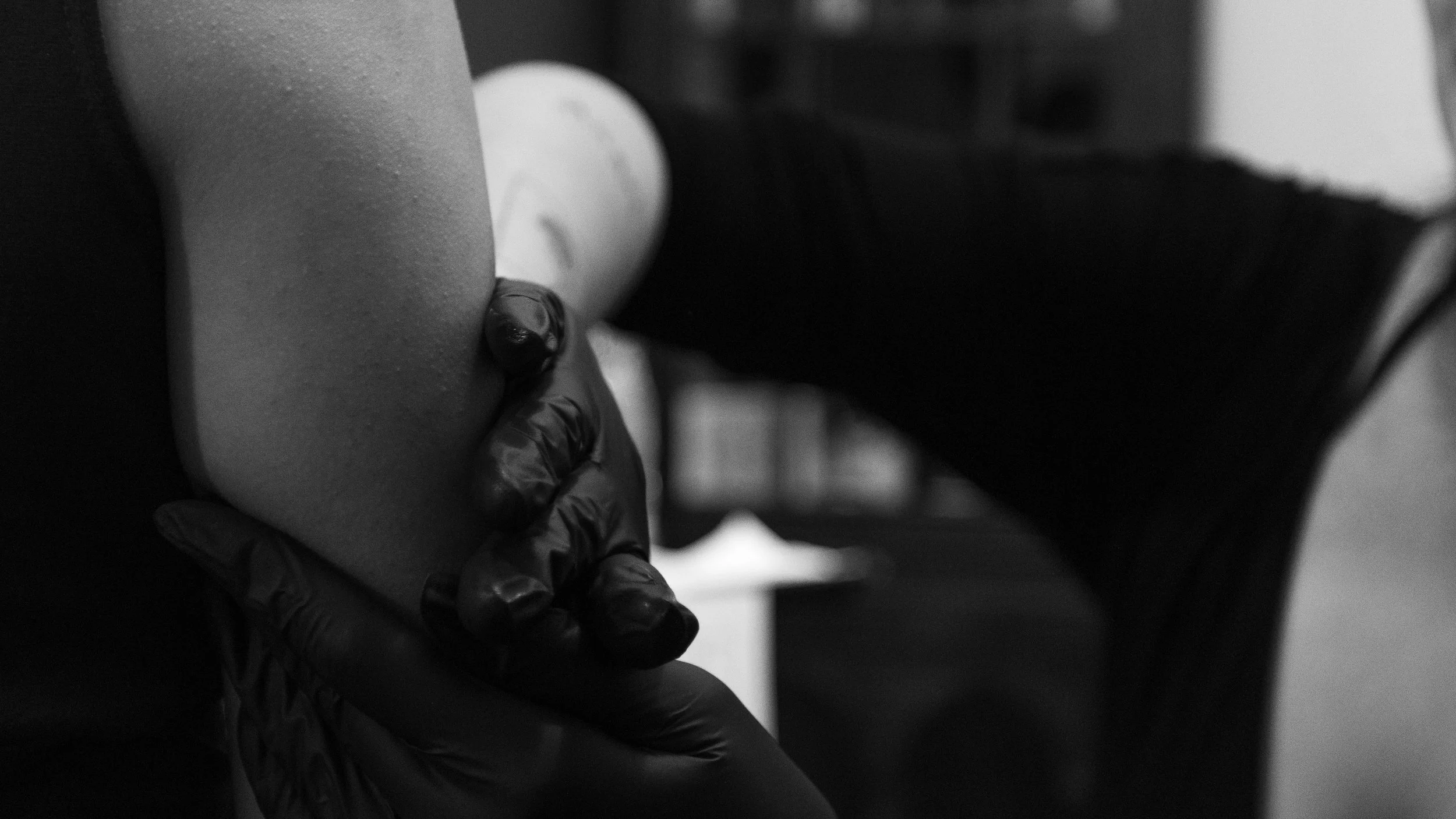 A close-up of a person wearing black gloves holding a tattoo machine, preparing to tattoo a person's arm in a tattoo studio.