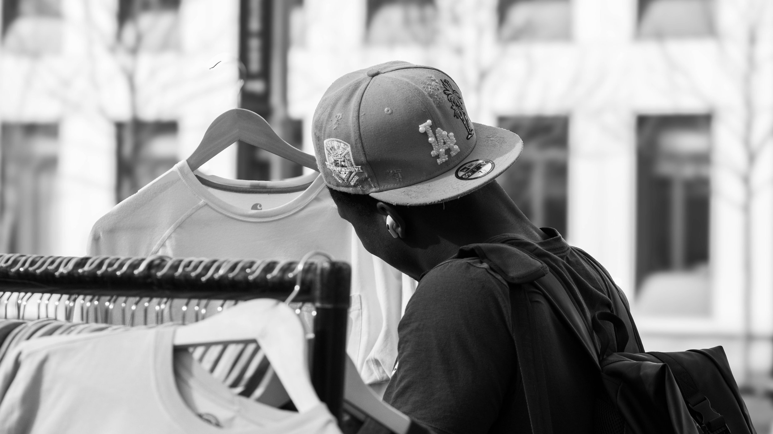A person with a backpack shopping at a clothing store. They are looking at t-shirts on a rack, wearing a cap with patches and a graphic on the front, and an earpiece.