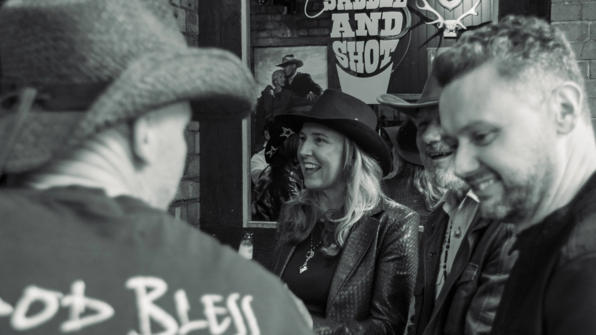People gathered in a bar with a cowboy-themed decor, engaging in conversation and smiling, with a sign in the background that reads 'SHADES OF SHOOT.'