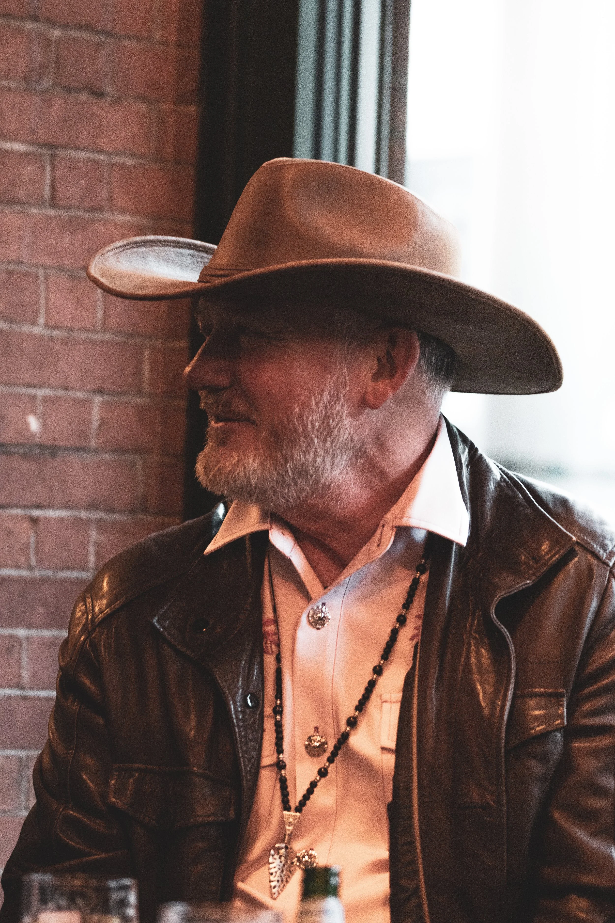 A man with a beard and glasses wearing a brown cowboy hat, leather jacket, and jewelry, sitting against a brick wall.