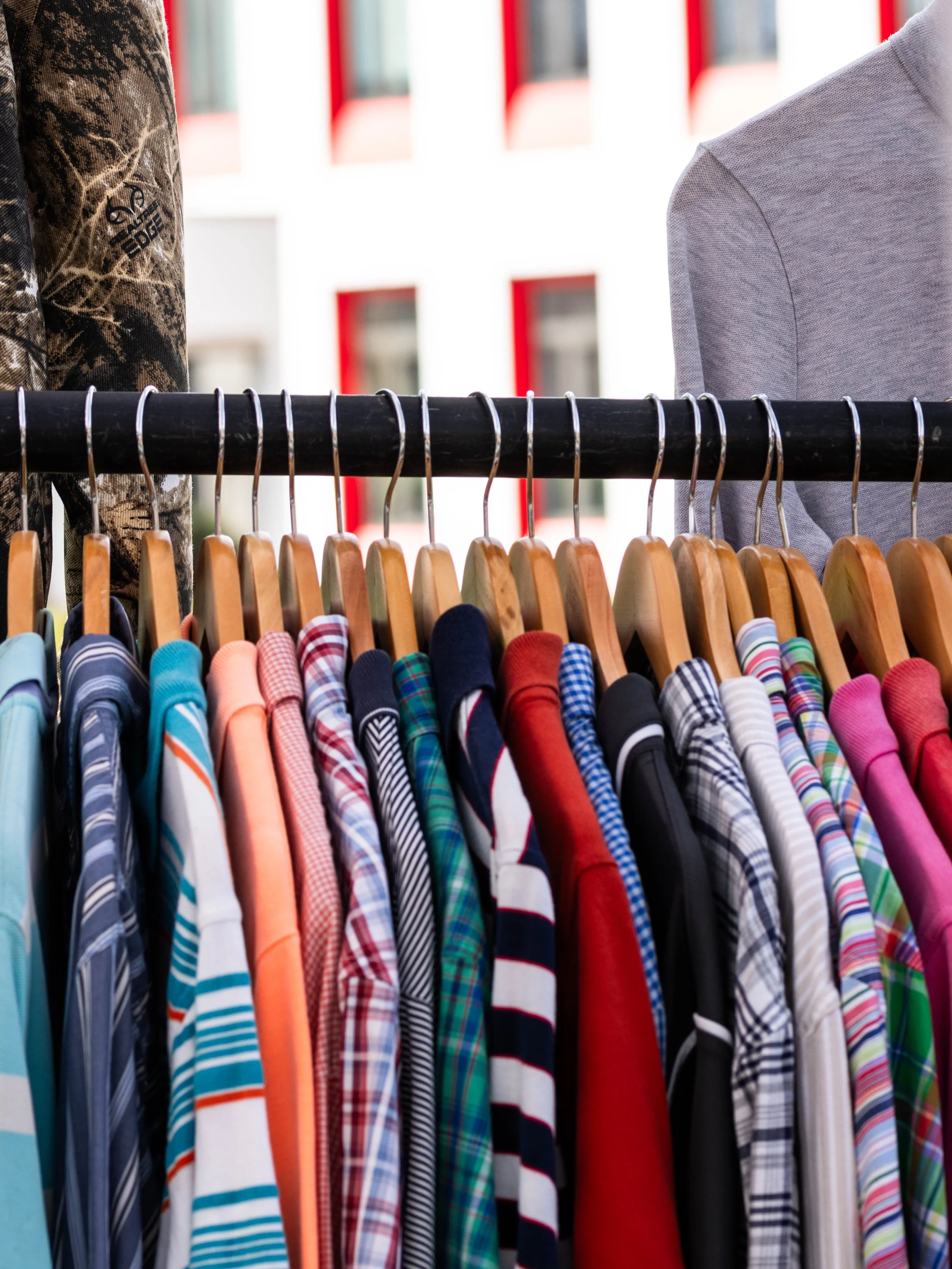 A row of colorful striped and plaid men's shirts on hangers at an outdoor flea market, with a blurred background of buildings.