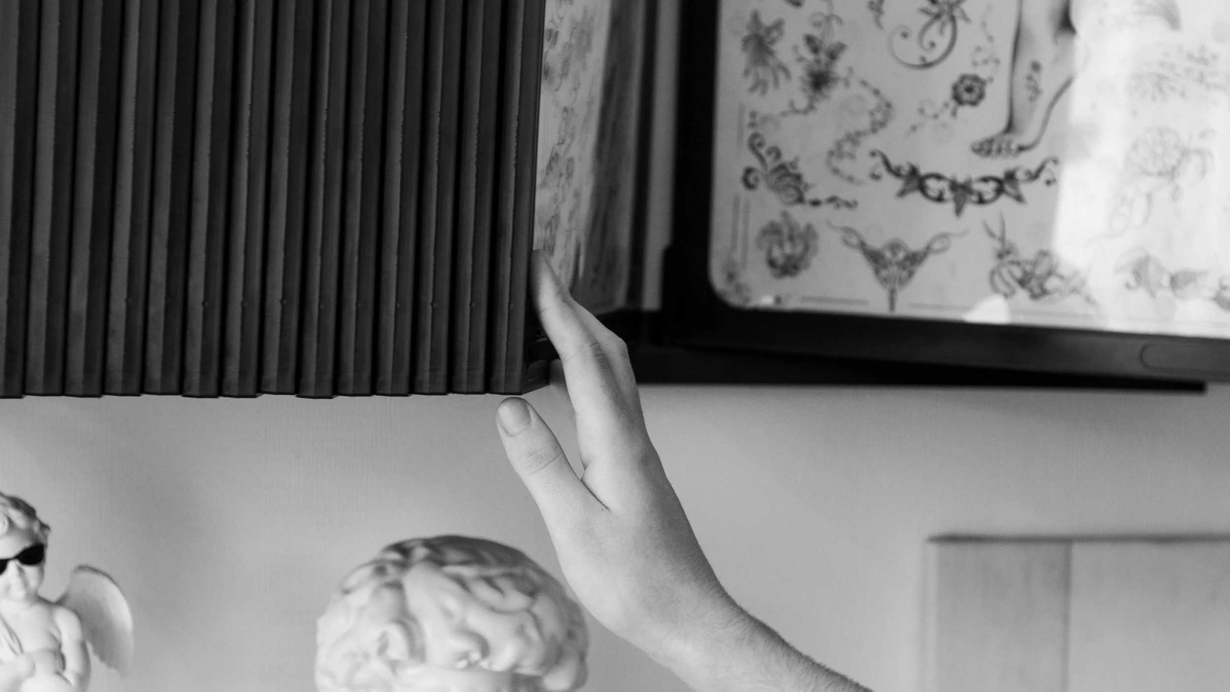 Black and white image showing a hand reaching out to touch a stack of black books. In the background, there is a framed artwork or photograph with floral and decorative patterns, and some small sculptures or figurines.