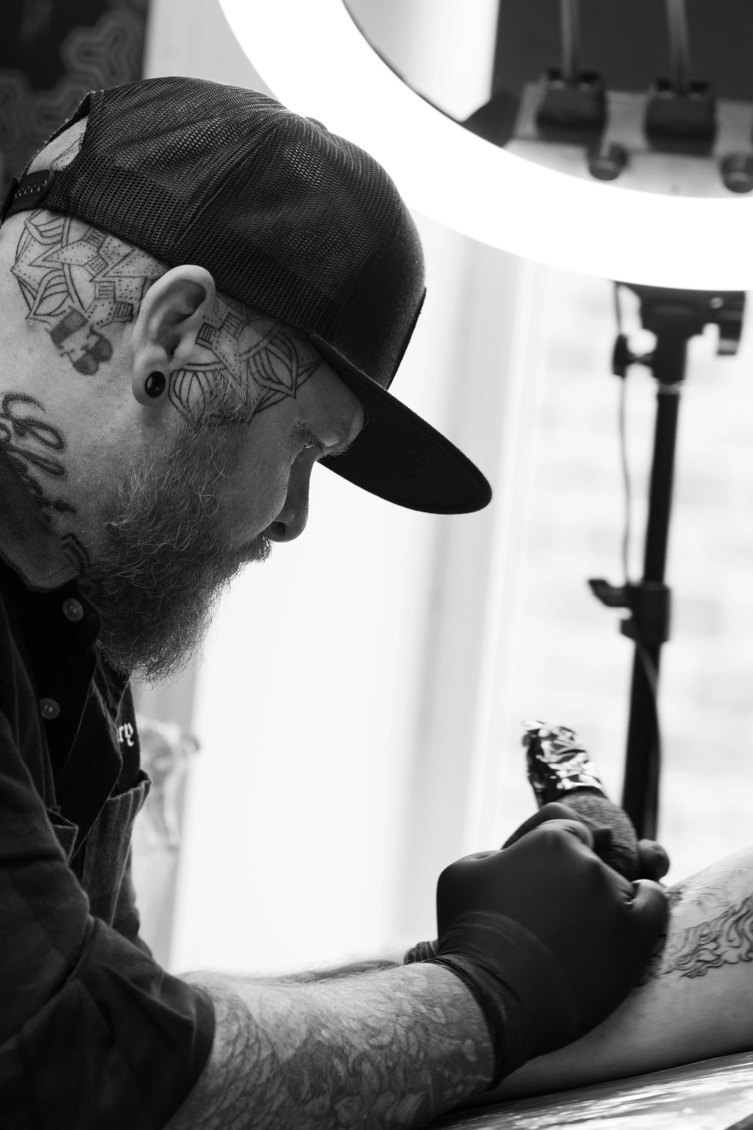 Black and white photo of a tattooed man with a beard and a baseball cap, sitting on a tattoo chair while getting a tattoo. There is a tattoo machine and a tattoo artist's gloved hand visible.