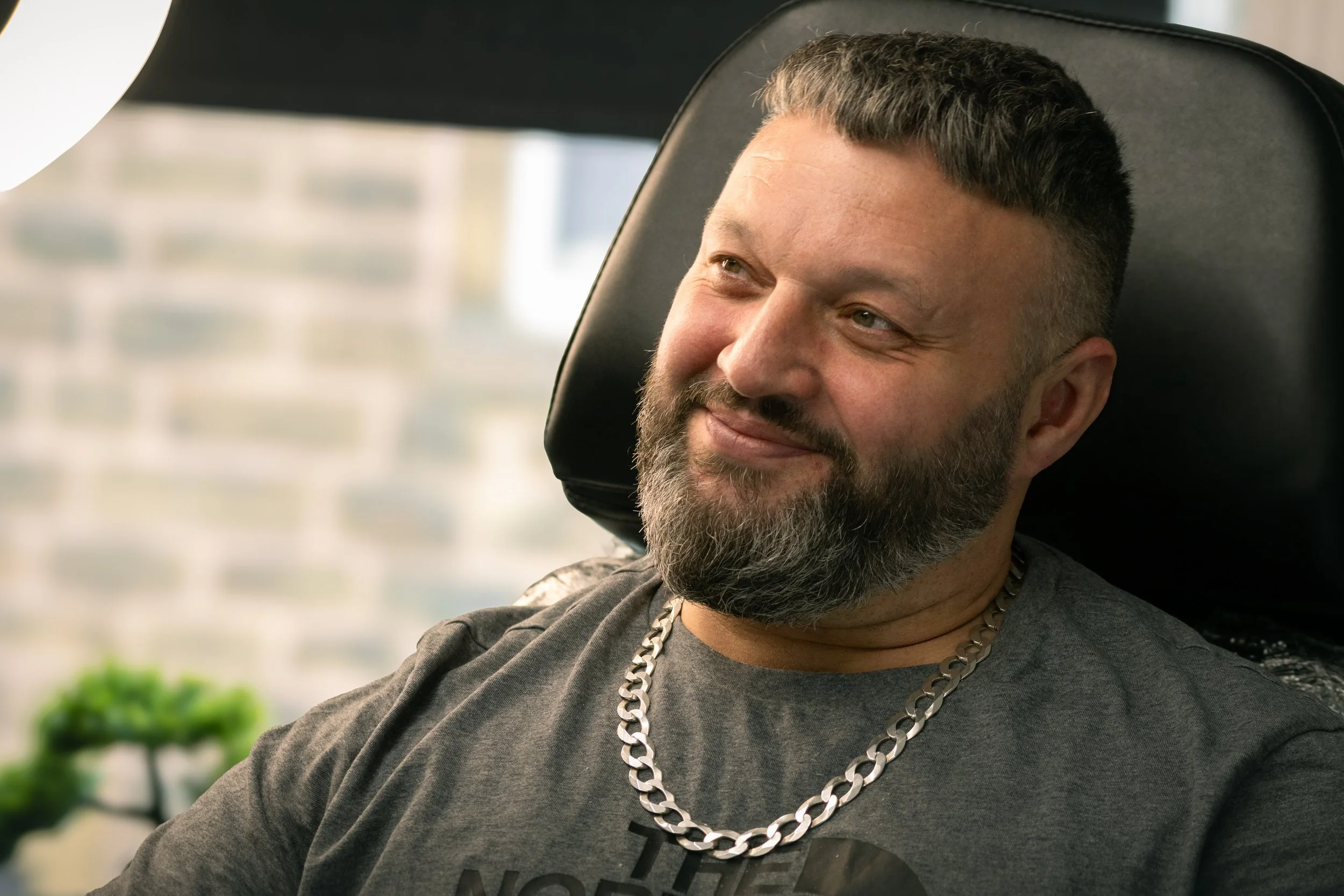 Close-up of a middle-aged man with short, dark hair and a beard, sitting in a chair with a black headrest, smiling and looking to the side, wearing a gray shirt and a silver chain necklace, in a brightly lit indoor setting.