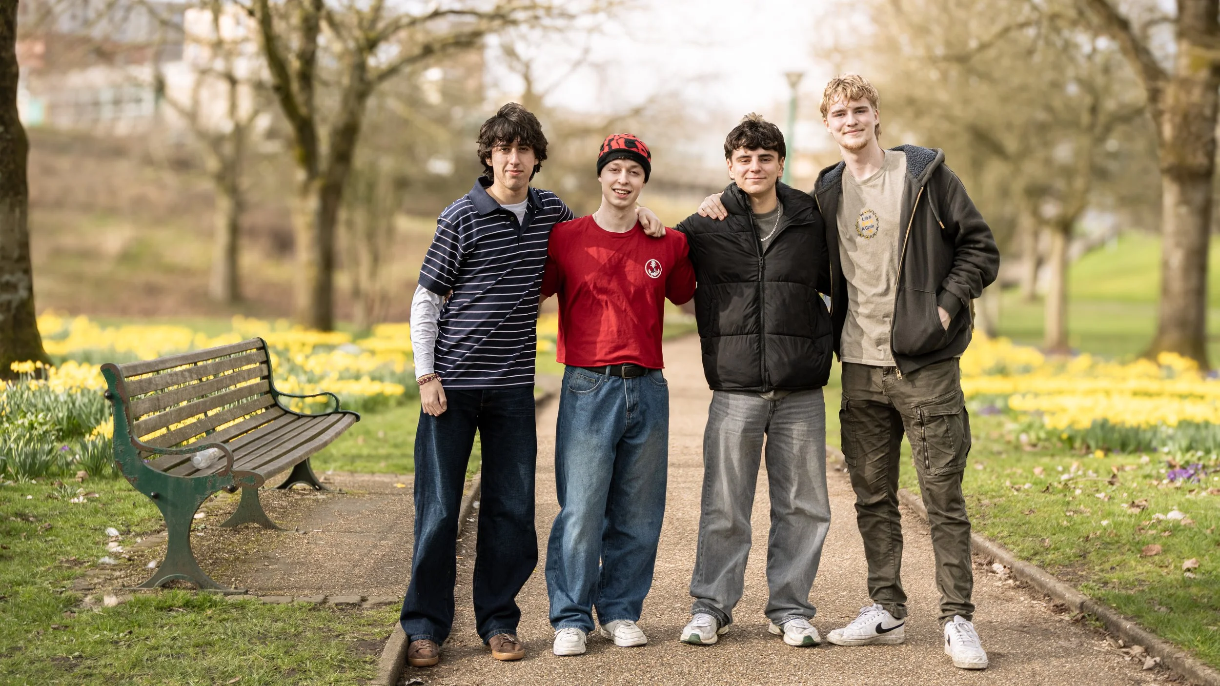Four teenage boys standing arm in arm on a park trail, with trees and yellow flowers in the background.