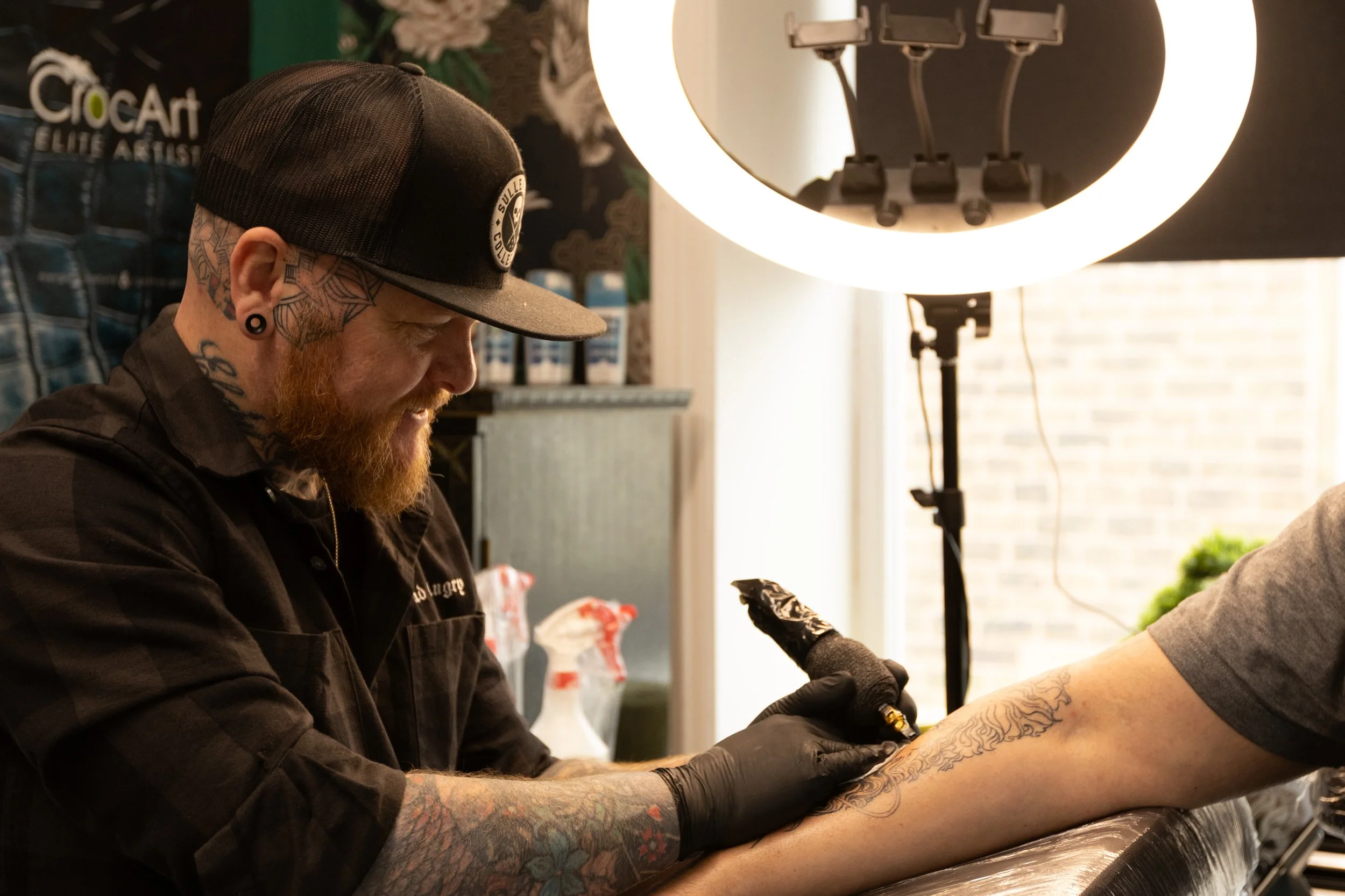 A man with tattoos and a beard, wearing a black trucker hat and black gloves, is getting a tattoo on his arm in a tattoo studio. There is a ring light behind him, and a manicurist is holding his arm.