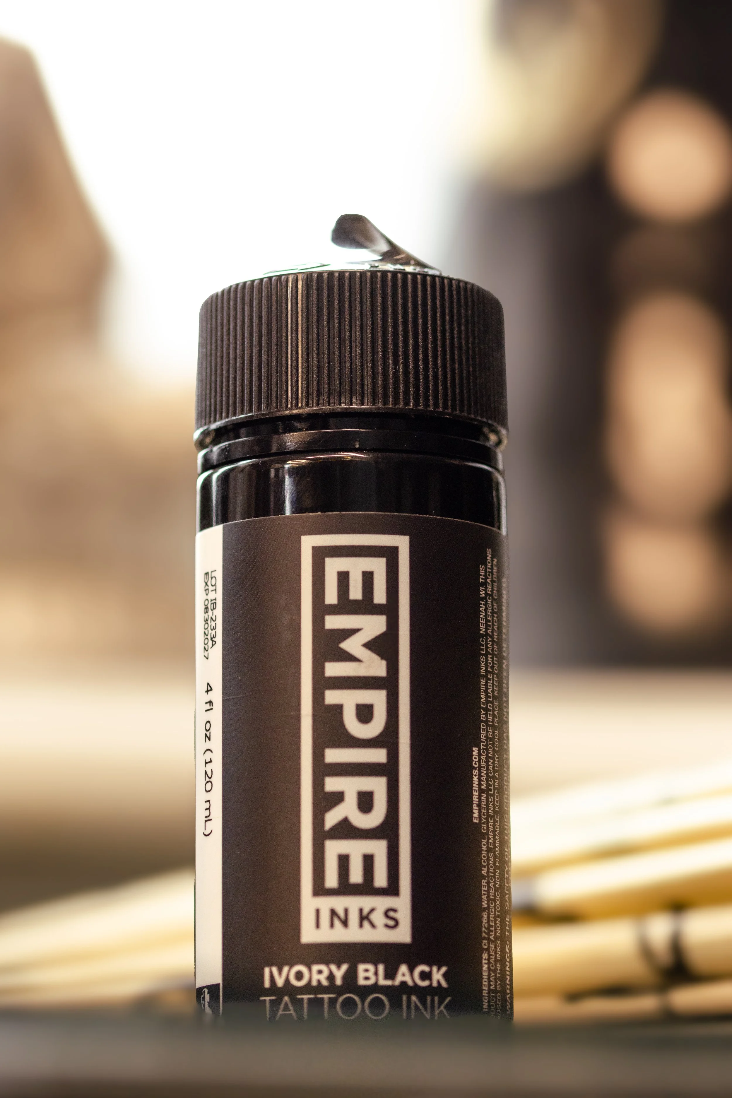 A black and white tattoo ink bottle labeled 'IVORY BLACK' with a black flip cap, placed on a wooden surface with blurred objects in the background.