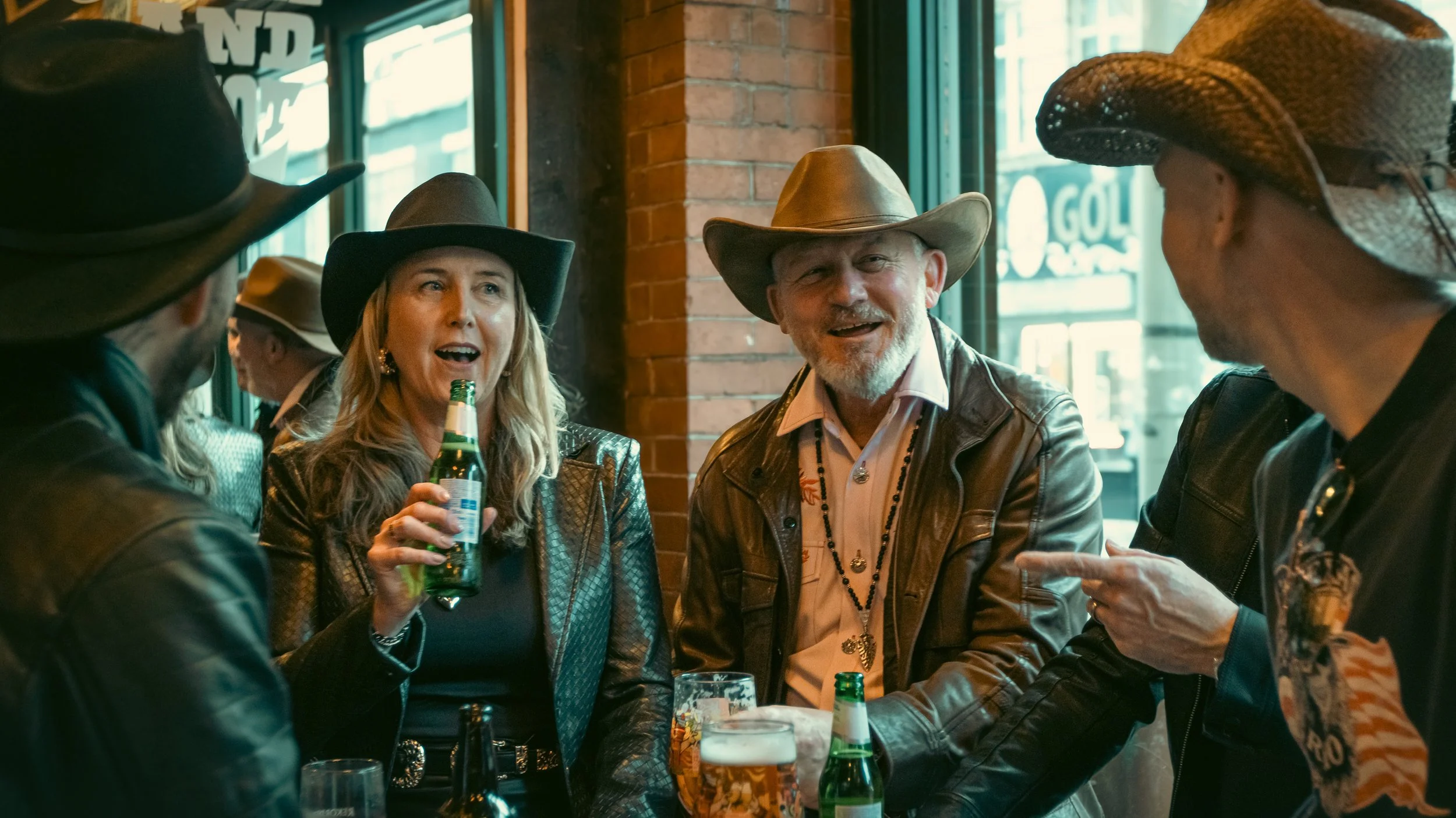 A group of people dressed in cowboy hats and western attire socializing at a bar. The woman in the center is holding a beer bottle and talking, while the men around her are engaged in conversation.