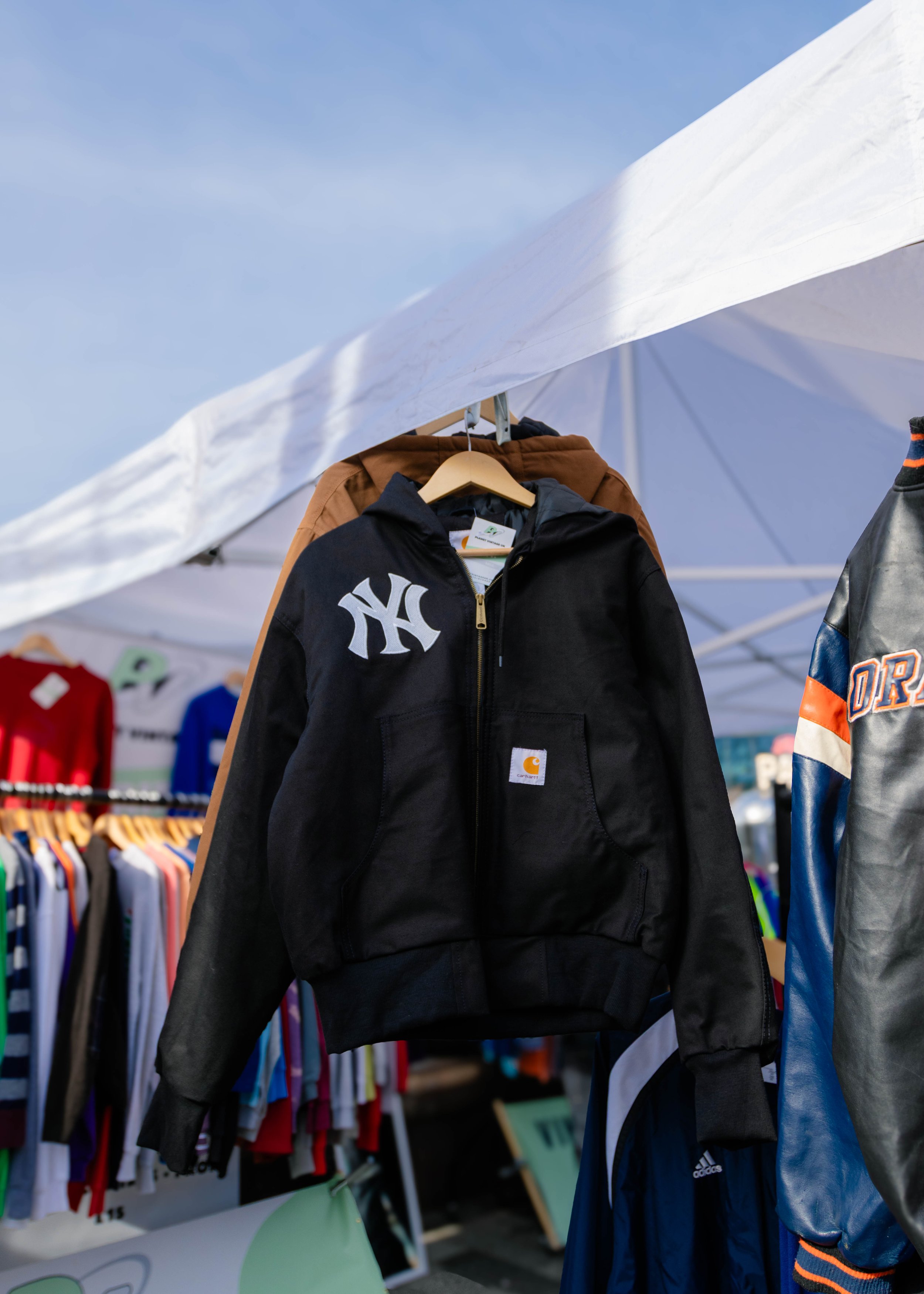 Clothing display at a retail store with a black Carhartt hoodie with a New York Yankees logo hanging in front, surrounded by other jackets, shirts, and sports apparel.