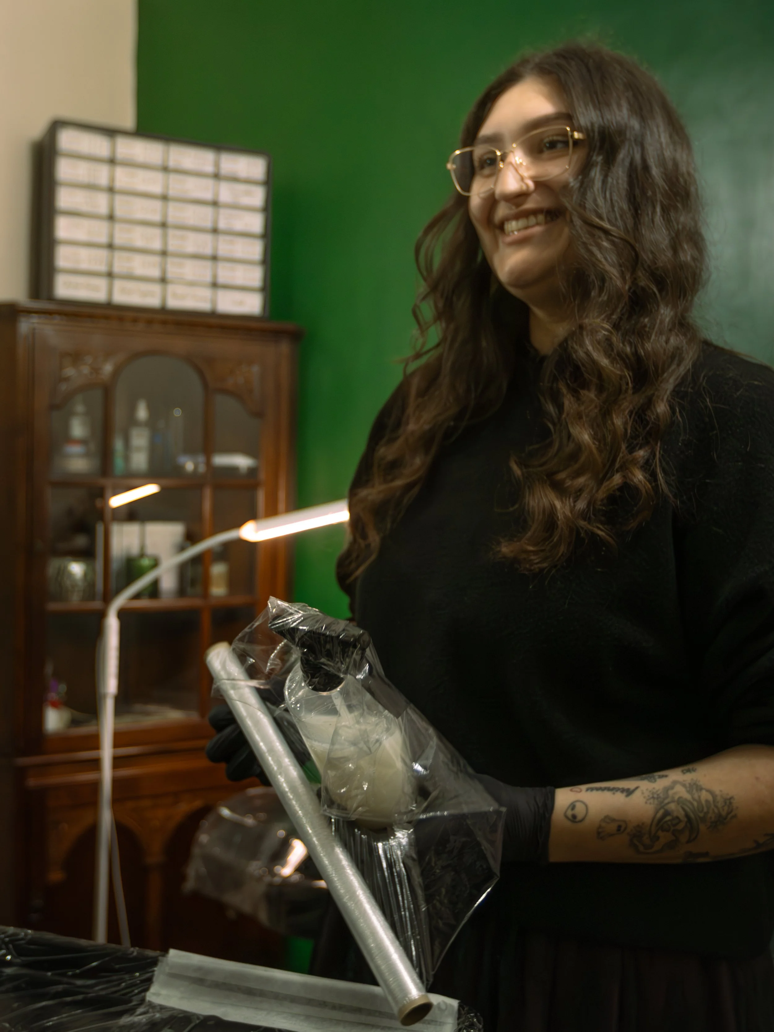 A smiling young woman with wavy, shoulder-length hair and glasses, wearing a black shirt, standing indoors near a green wall, holding a plastic bag with what appears to be black items inside, with a tattooed arm and a tattooed wrist visible.