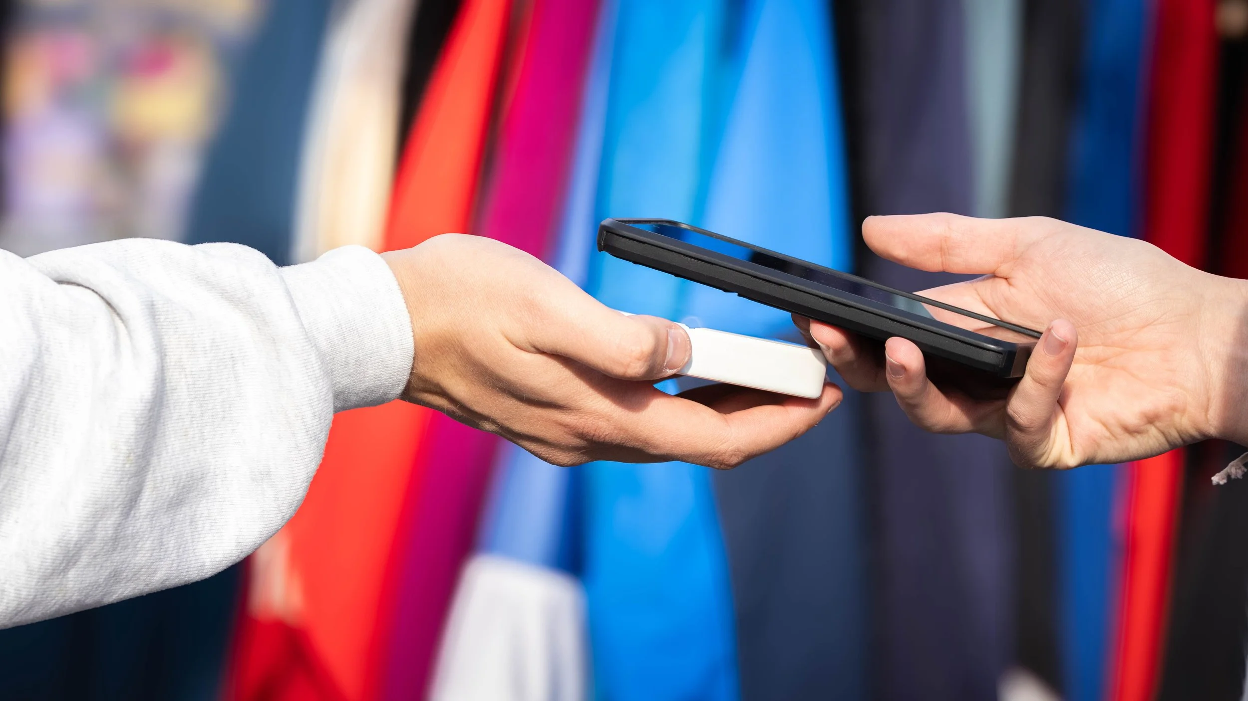 Close-up of one person handing a credit card and smartphone to another person against a background of colorful clothing racks.