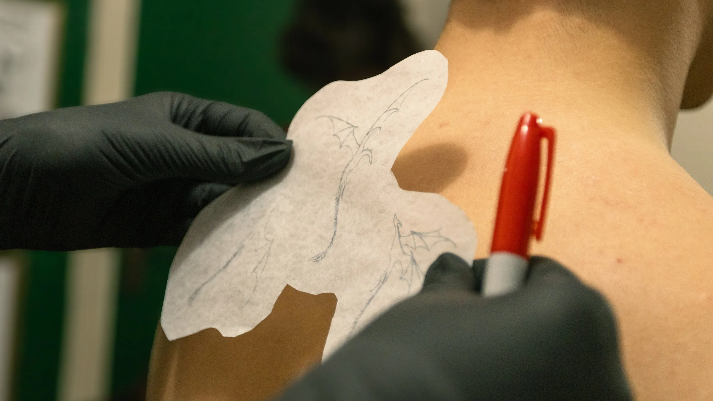 A person wearing black gloves is drawing a dragon tattoo outline on a person's arm using a red marker, with a paper stencil nearby.
