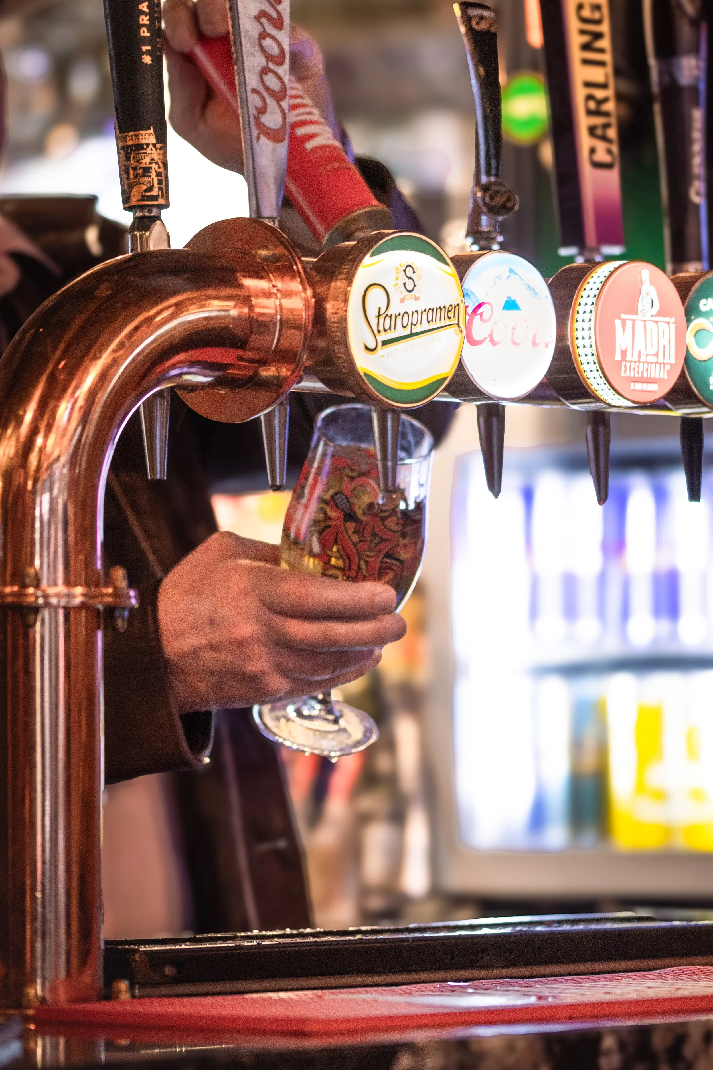 Close-up of a bartender filling a glass at a bar with beer taps, featuring brands such as Staropramen, Coca-Cola, and Madri. The bartender's hand is visible, holding the glass under the tap as foam forms at the top.