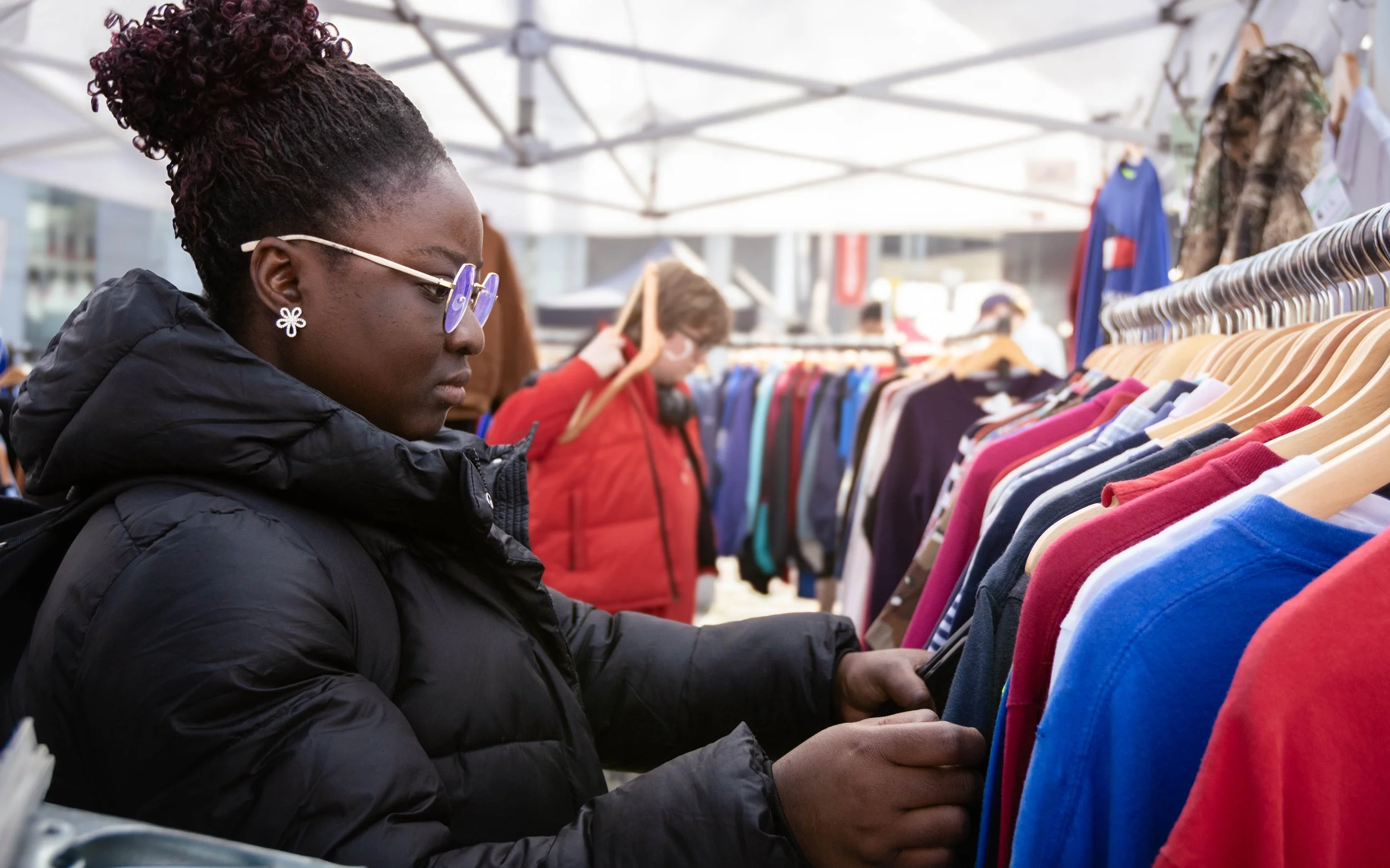 Woman with curly hair tied up, wearing glasses and a black jacket, shopping for clothes at an outdoor market with racks of colorful shirts.