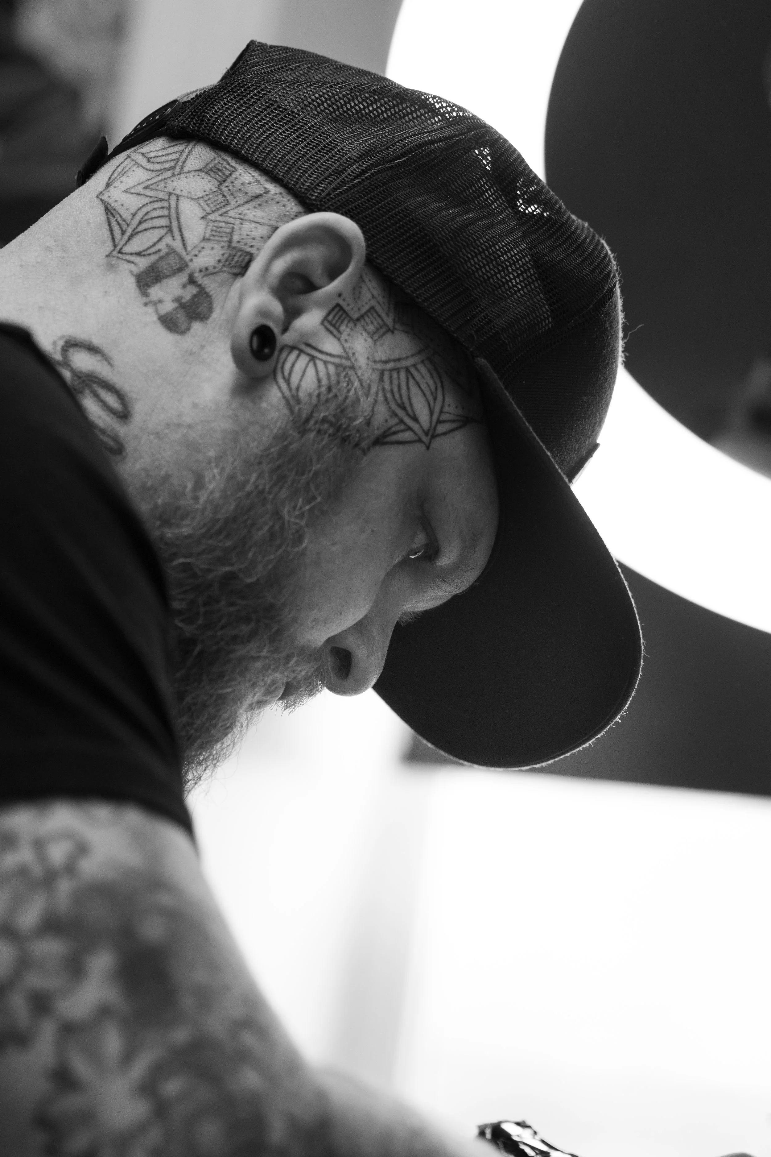 Close-up black and white photograph of a man with tattoos on his face, beard, and ear piercing, wearing a backwards baseball cap. The angle is from below, capturing his face partially in profile with a bright light or reflector in the background.