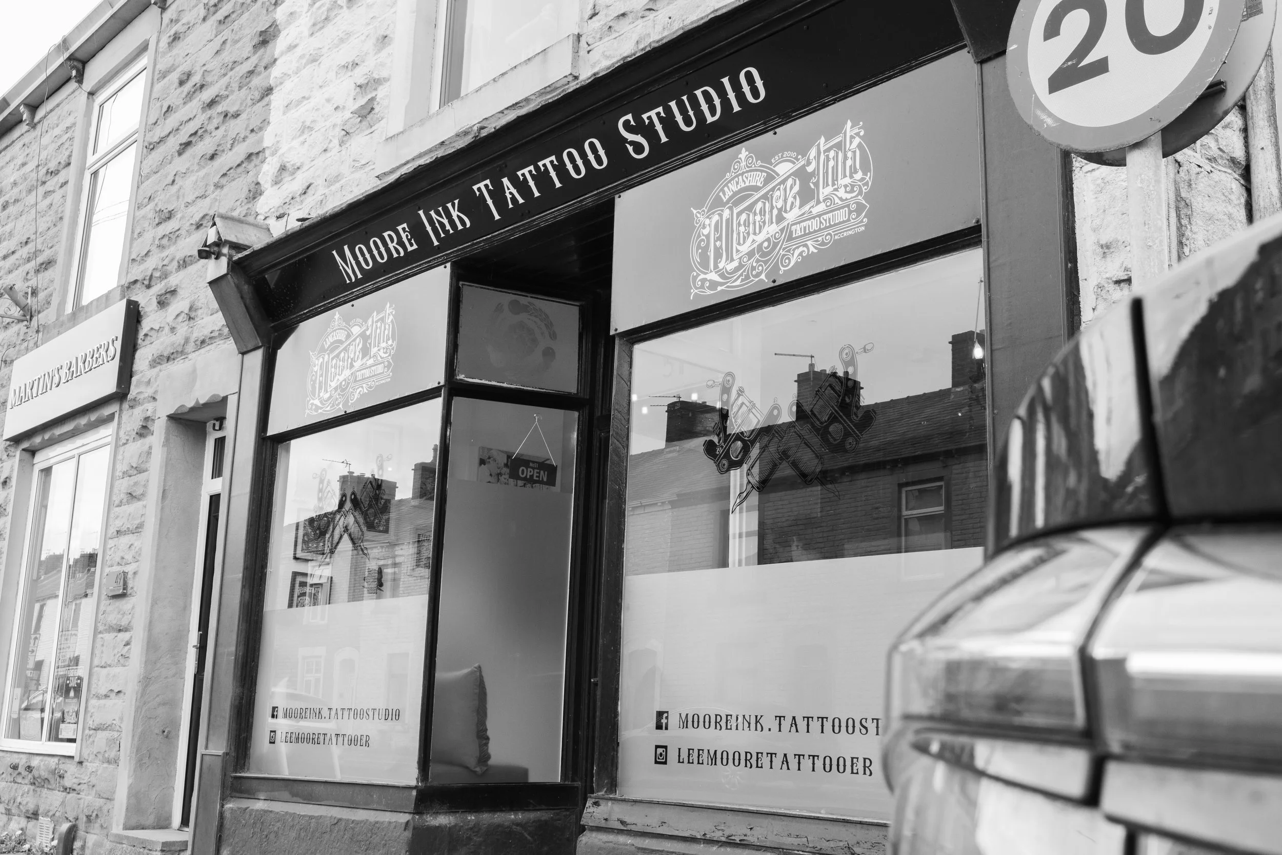 Black and white photo of a storefront with a sign reading 'Moore Ink Tattoo Studio,' with contact details and social media icons on the glass window.