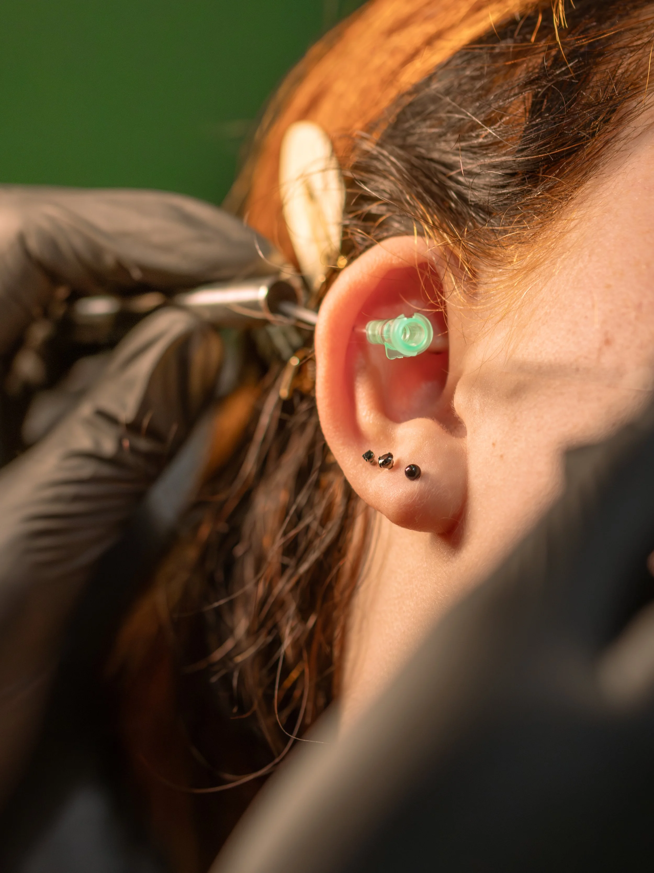 A person getting a hearing aid fitted in their ear, with a close-up of the ear and hair, and the fitting device and gloved hand visible.