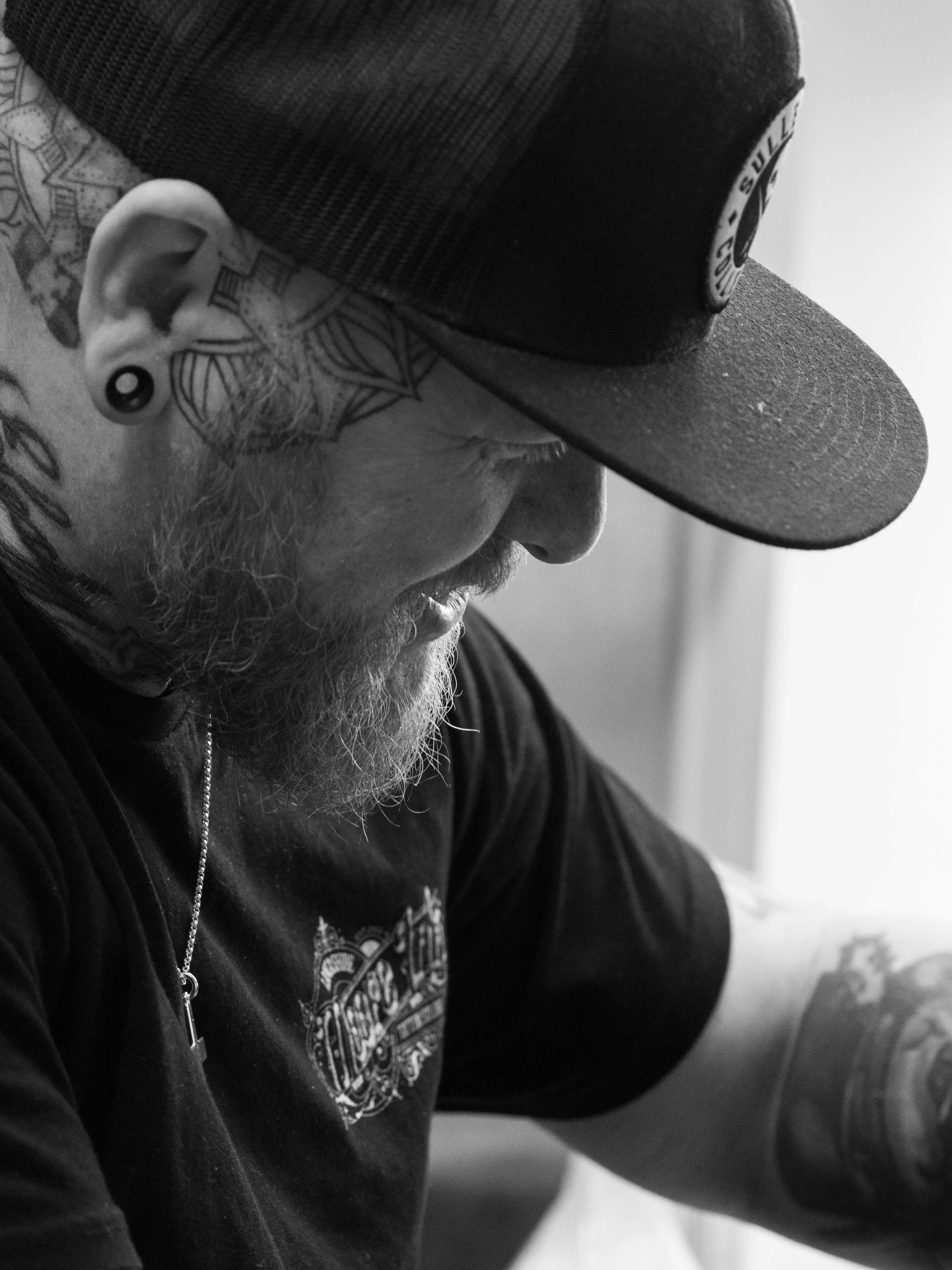 Close-up black-and-white photo of a man wearing a baseball cap, tattoos on neck and ear, and a chain necklace, with a contemplative expression.
