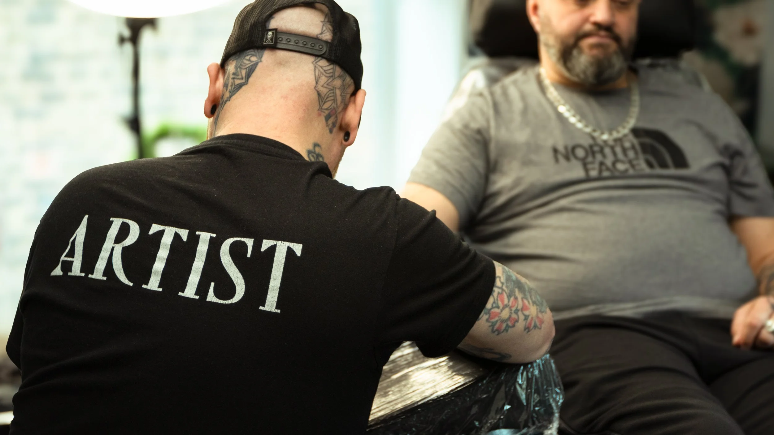 Two men sitting side by side, with the man on the left wearing a black cap backward and a black shirt with the word 'ARTIST' visible, tattoos on his neck and arms, and the man on the right wearing a gray North Face shirt, a chain necklace, and having