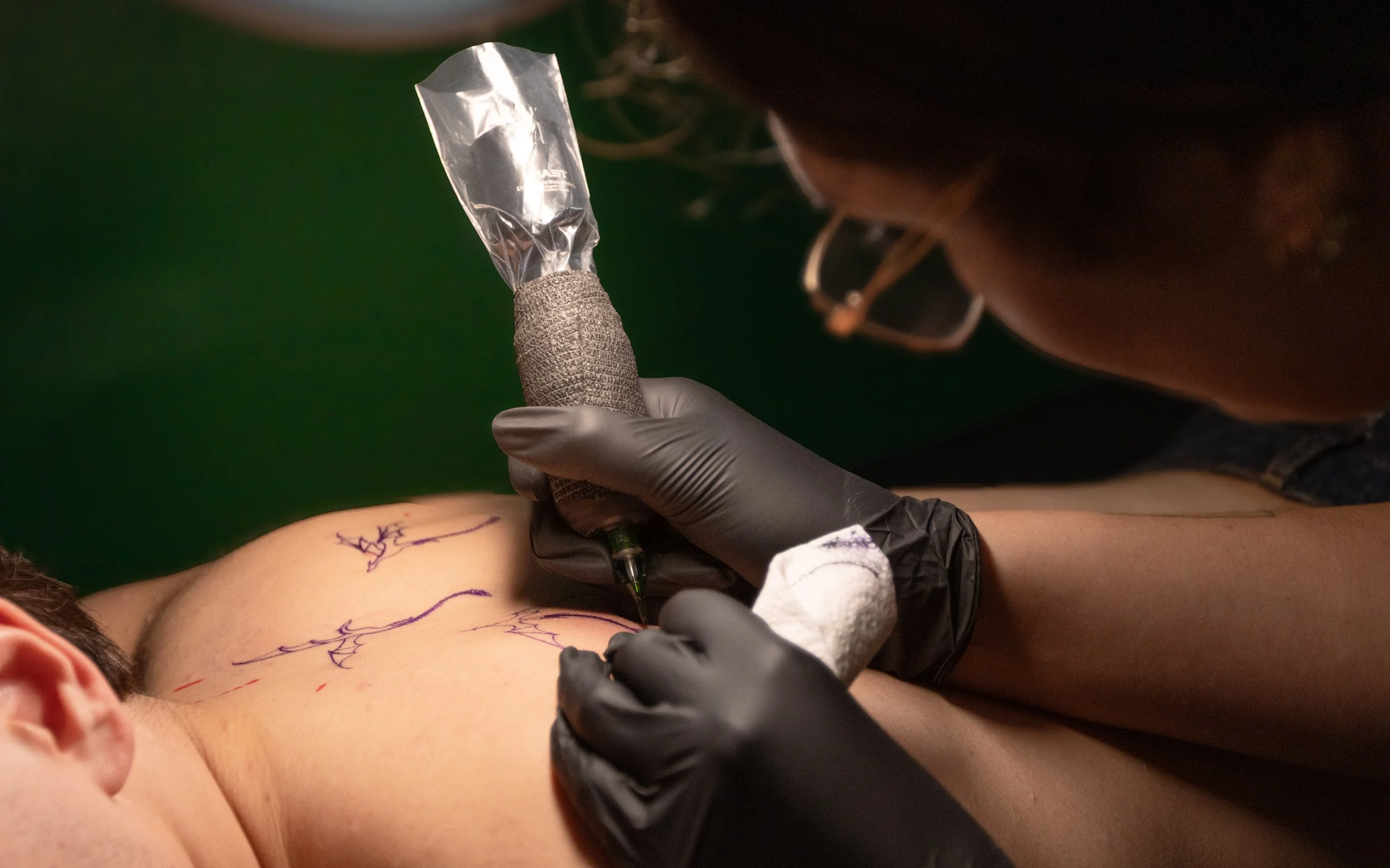 Tattoo artist using a tattoo machine to ink a design on a person's forearm, with visible outlines and sketches on the skin.