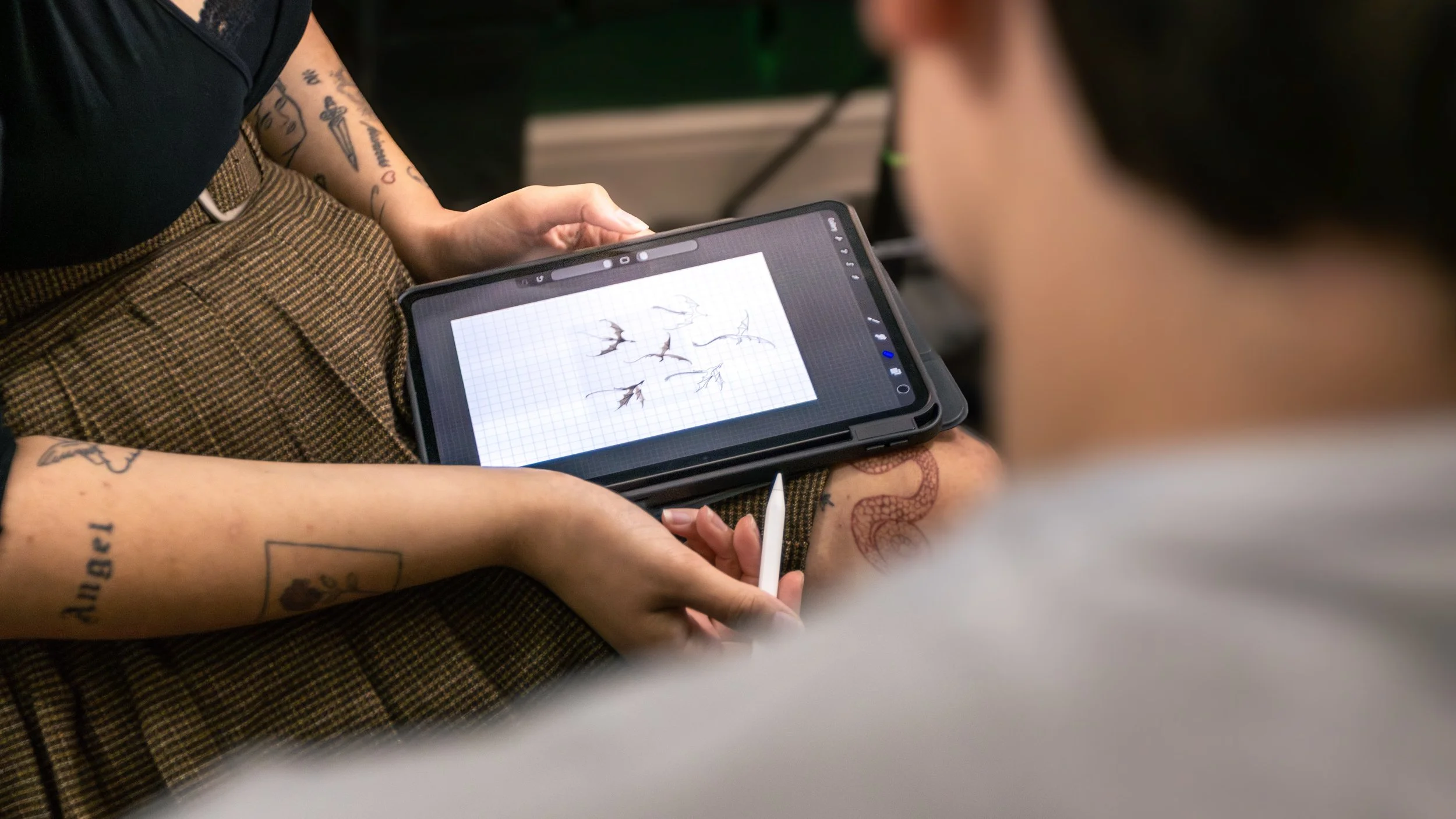Person holding a tablet with a drawing of branches and leaves on graph paper screen, while another person appears nearby.