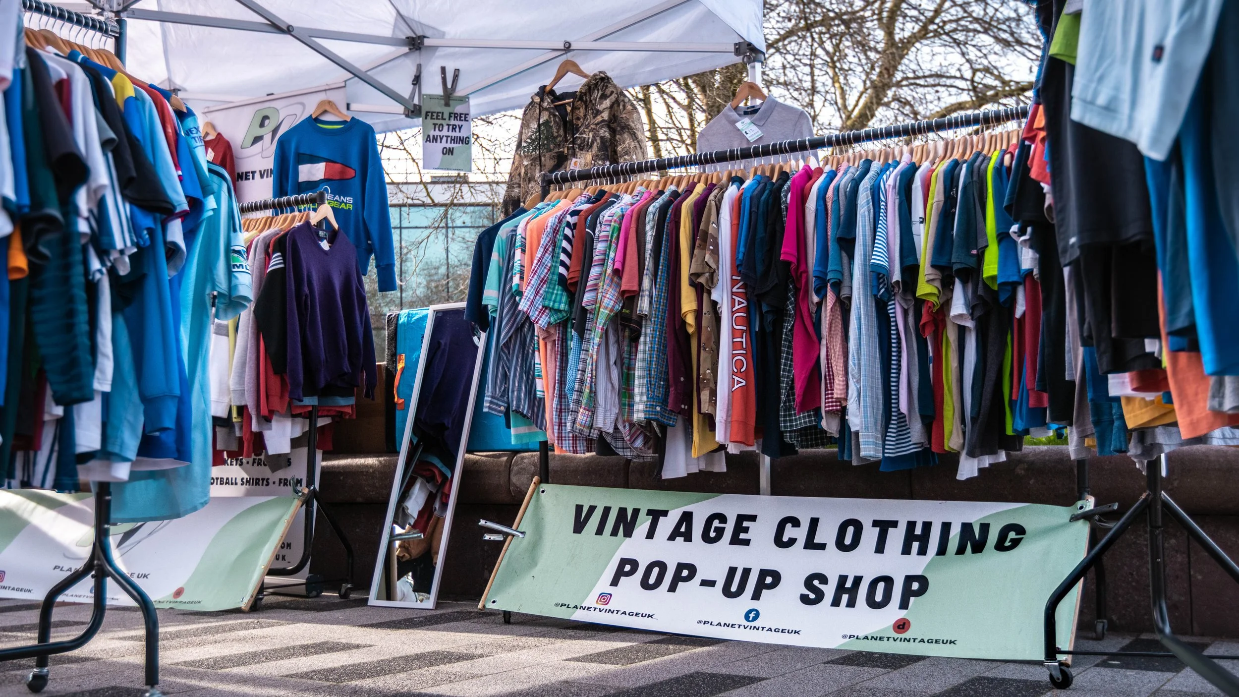 Vintage clothing pop-up shop with clothing racks filled with colorful shirts, jackets, and sweaters under a tent. There is a sign promoting the shop as a vintage clothing popup. Trees are visible in the background.