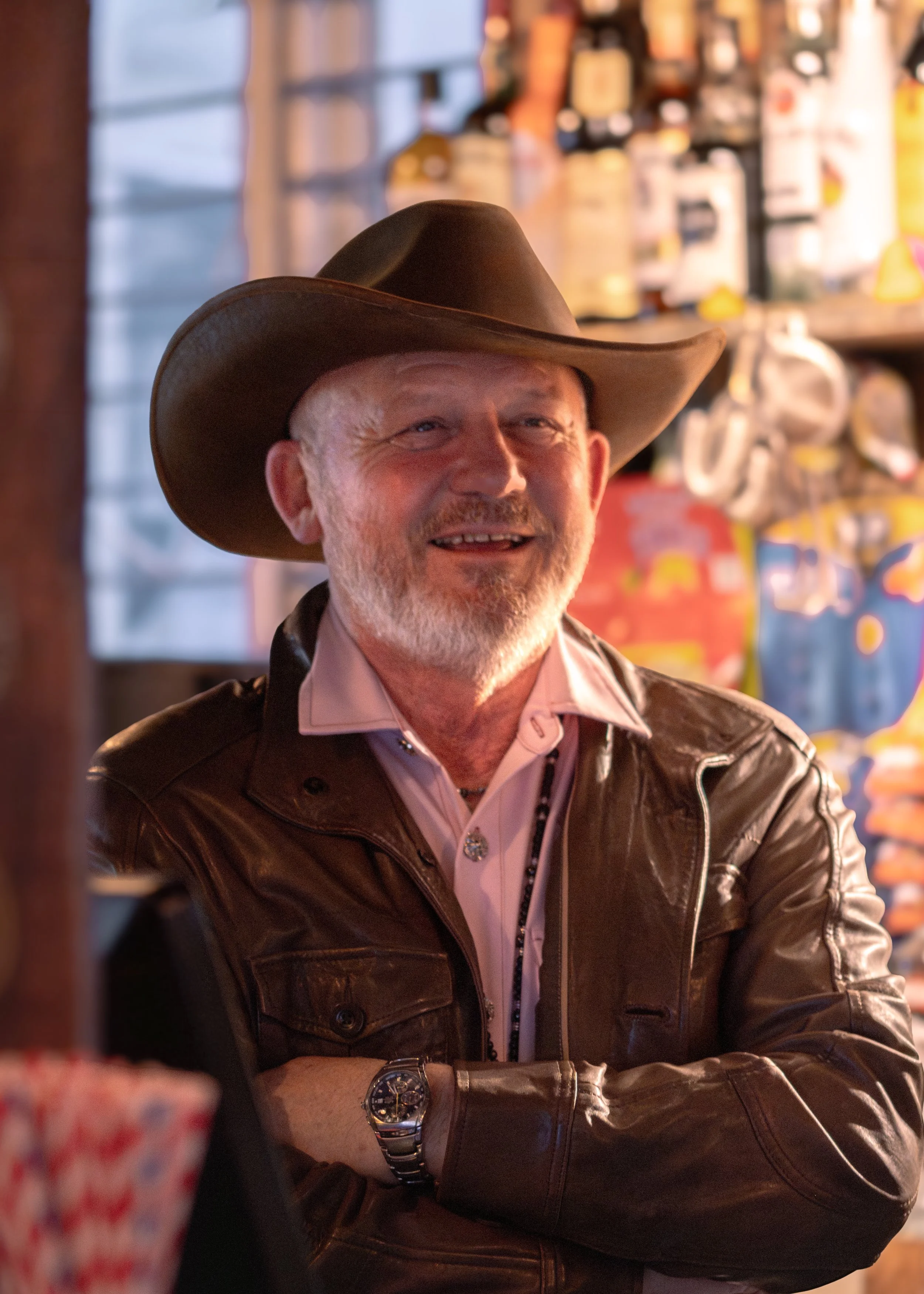 A man wearing a cowboy hat and leather jacket, smiling with his arms crossed, sitting at a bar or restaurant with shelves of bottles and snacks in the background.