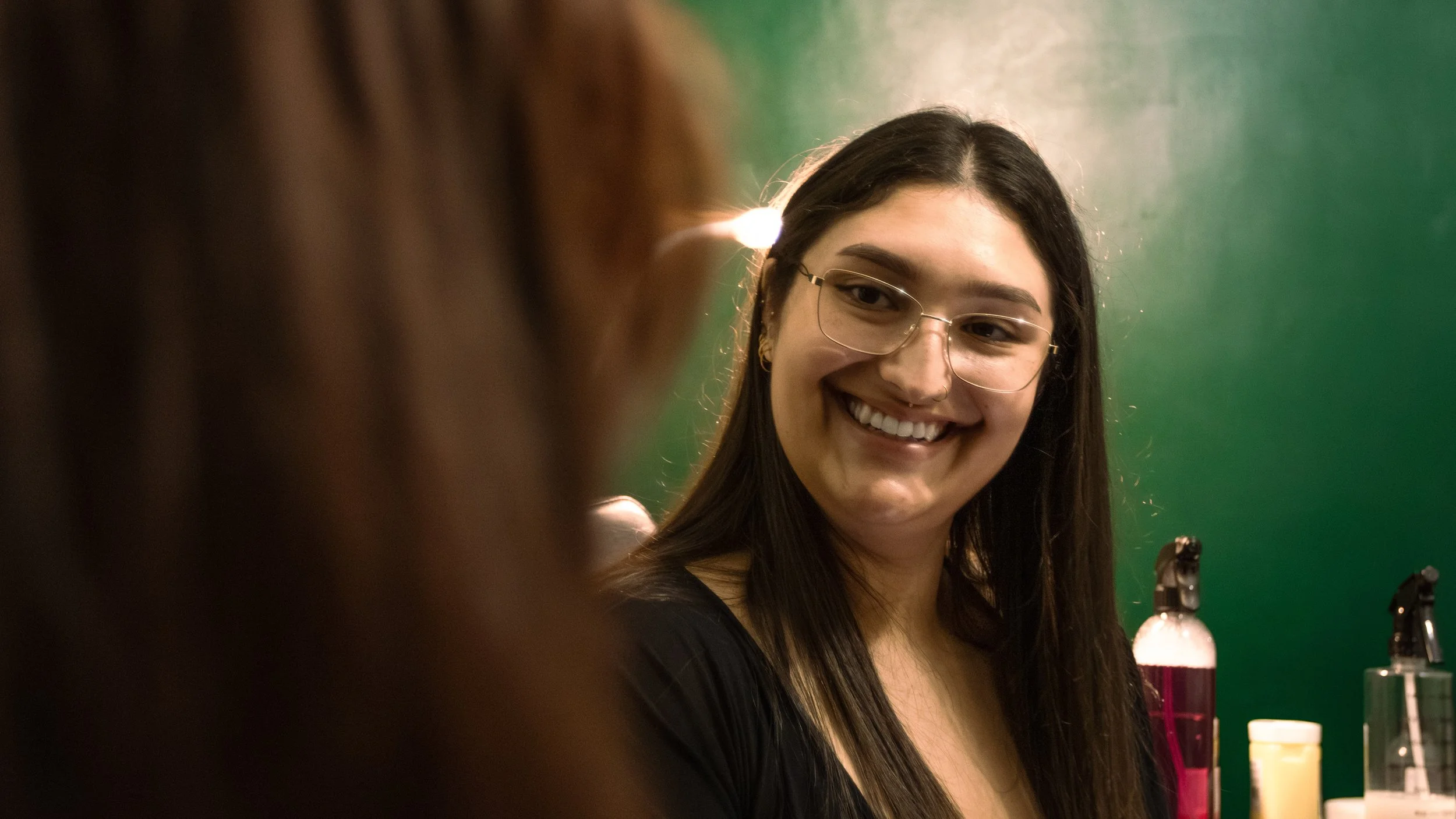 A young woman with long dark hair and glasses smiling while looking at someone in front of her. She is in a laboratory or science setting with colorful bottles and a green background.