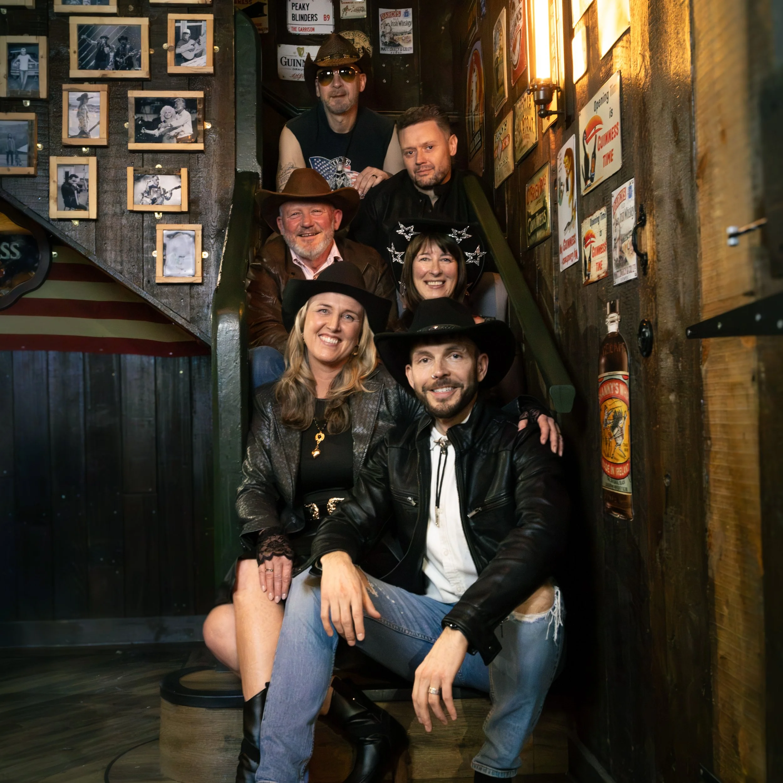 A group of seven people posing together in a bar or pub, with some wearing costumes like cowboy hats and witch hats. The background features a wall decorated with black and white photographs, posters, and signs related to Irish whiskey and Guinness.