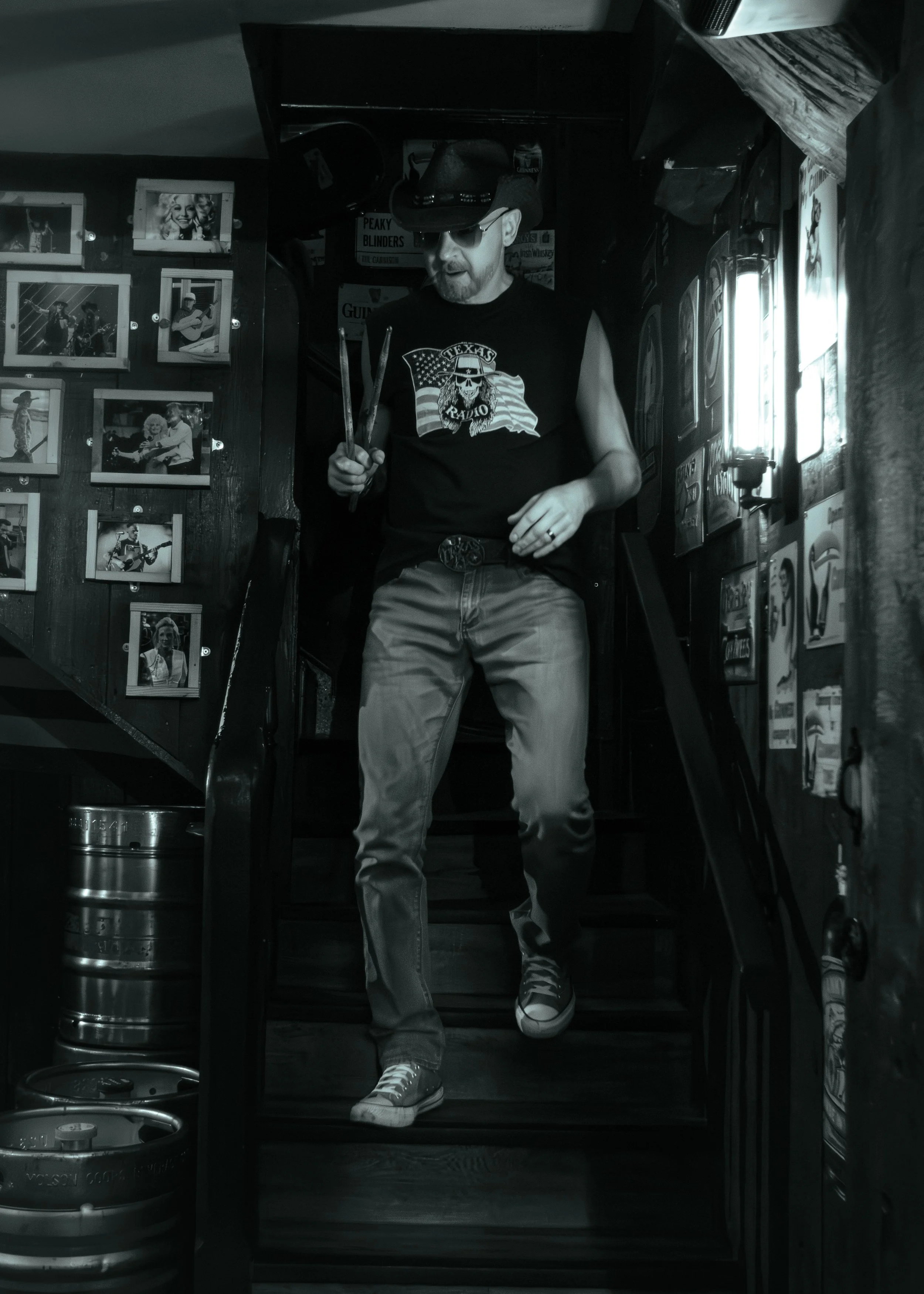 A man walking inside a bar, wearing a black sleeveless t-shirt with a skull graphic and the words 'Texas Radio', a black cap, sunglasses, and casual pants. He is holding a pair of chopsticks. The bar has framed photos on the wall and various signs, w