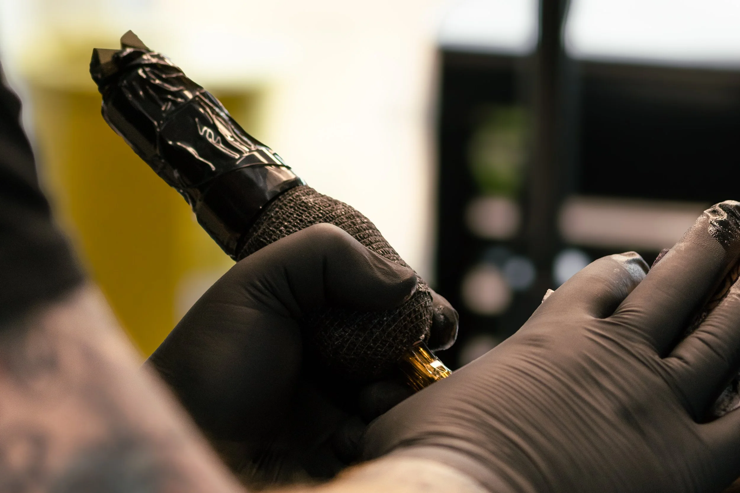 Close-up of a person wearing a glove, tattoo on their hand, and holding a tattoo machine, with a blurred background.