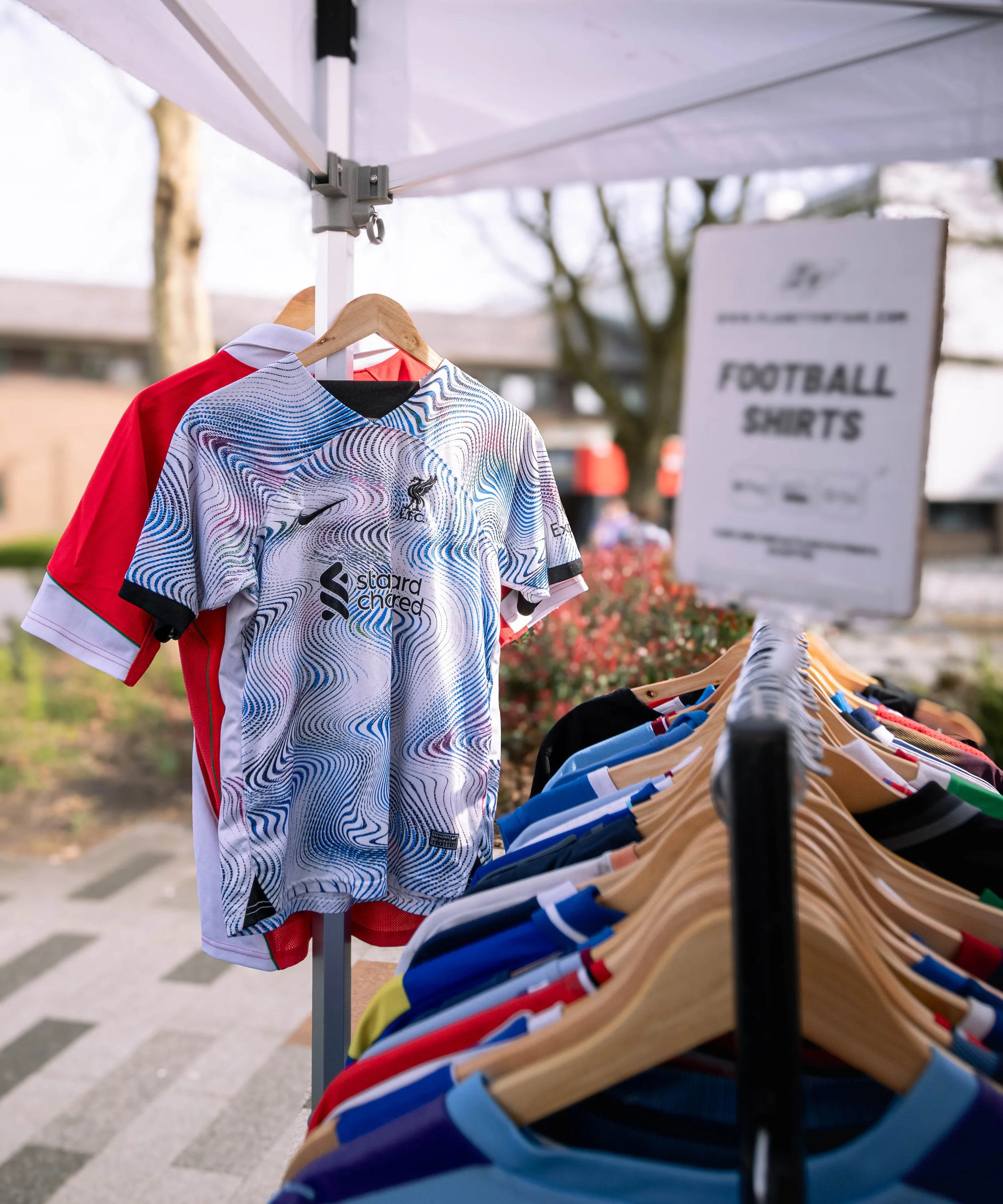 Clothing rack with colorful football jerseys displayed outside, under a white canopy, with a blurred background of trees and a sidewalk.