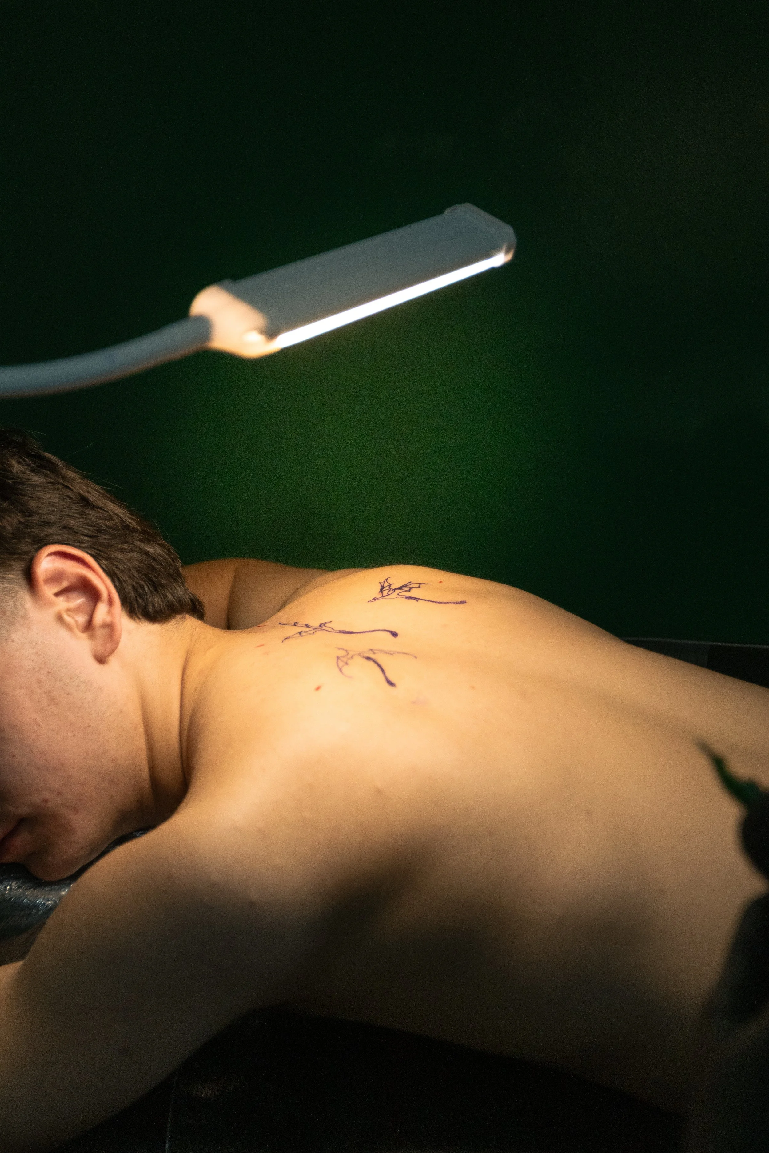 A person lying on their side, shirtless, with blue ink markings on their shoulder and upper arm, and a bright light directed at their skin, with a green background.