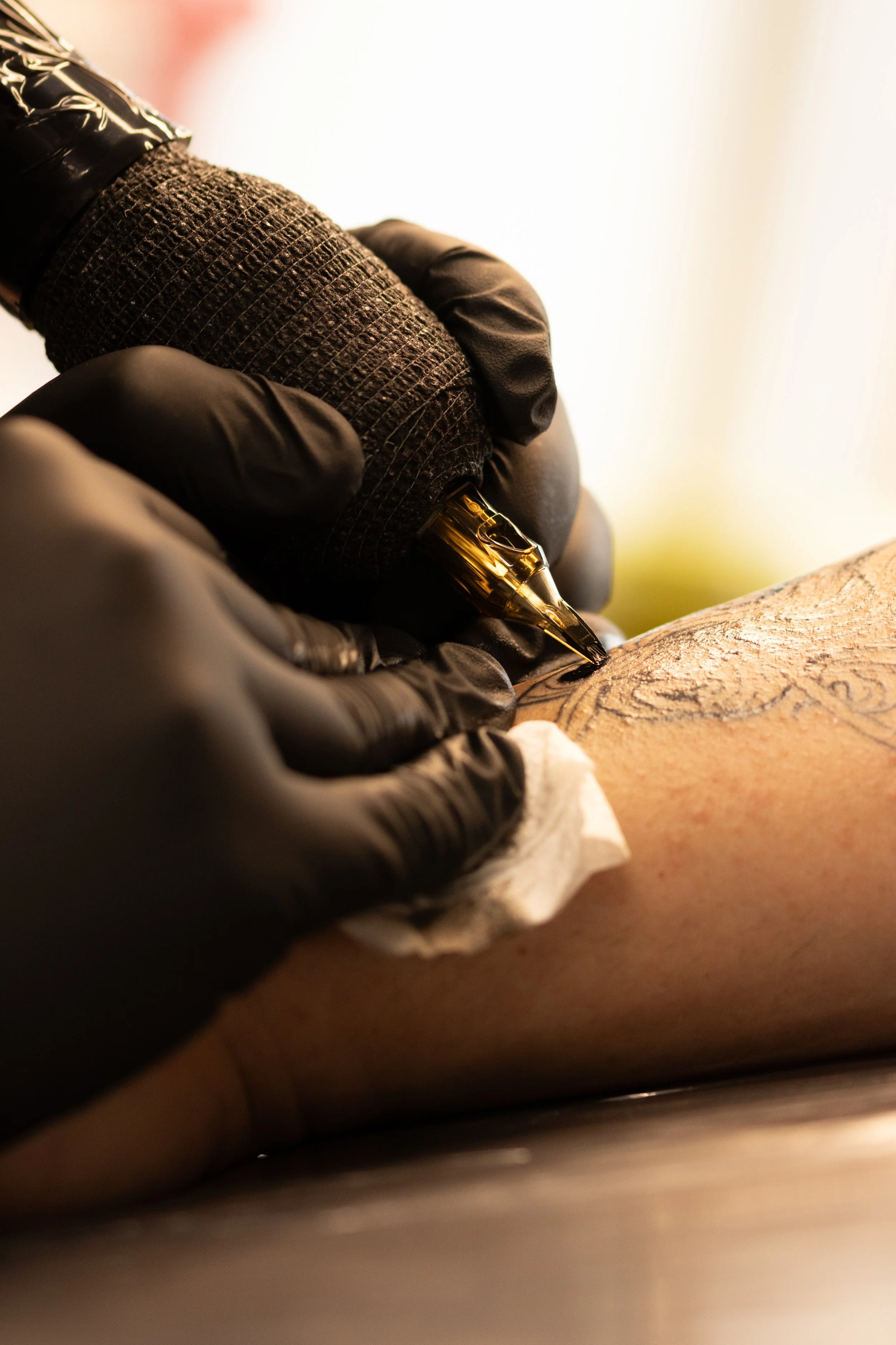 A person wearing black gloves tattooing a detailed design on someone's skin with a tattoo machine.