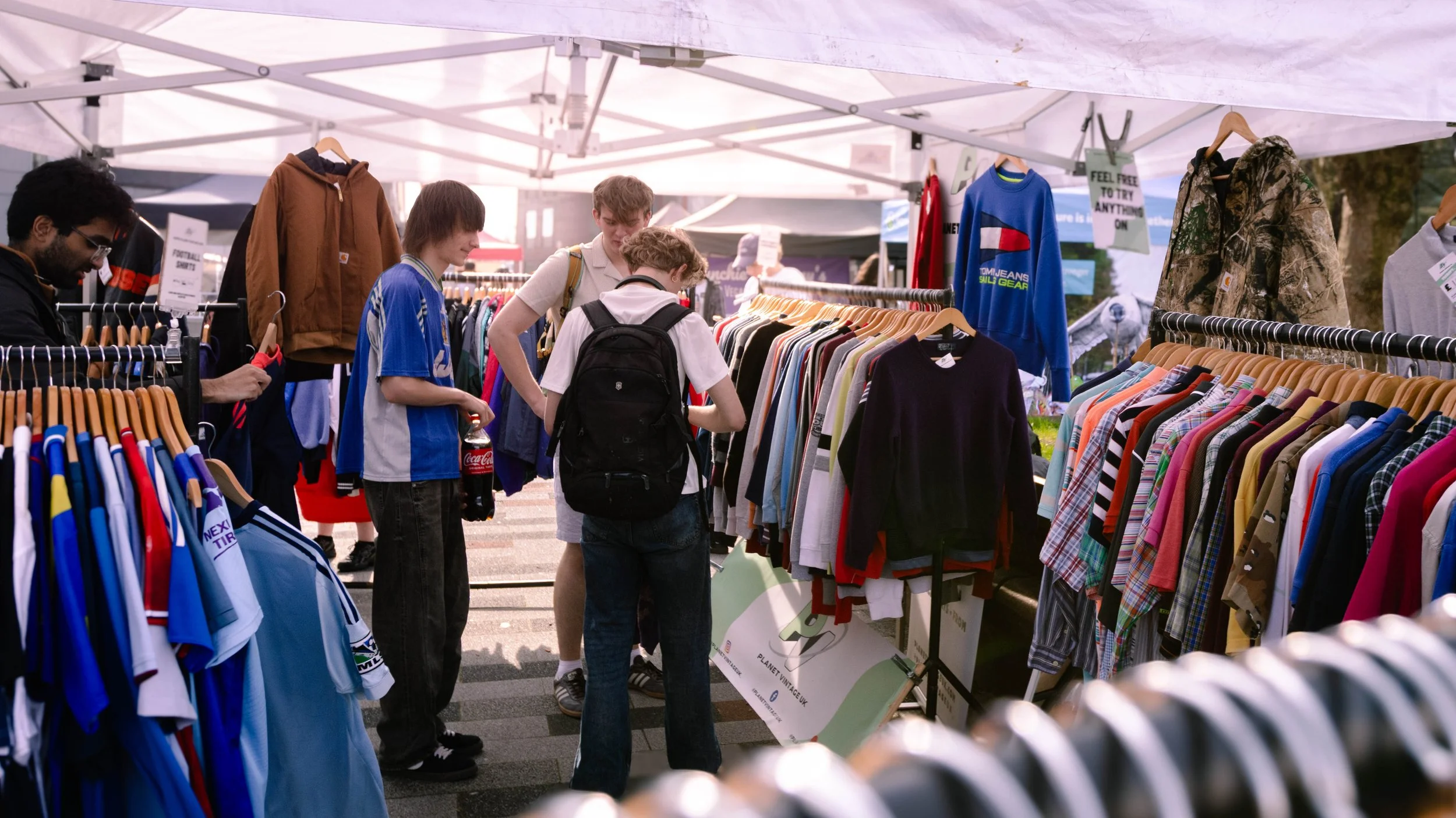 People shopping at an outdoor clothing market with racks of colorful clothes and jackets, under a white canopy.