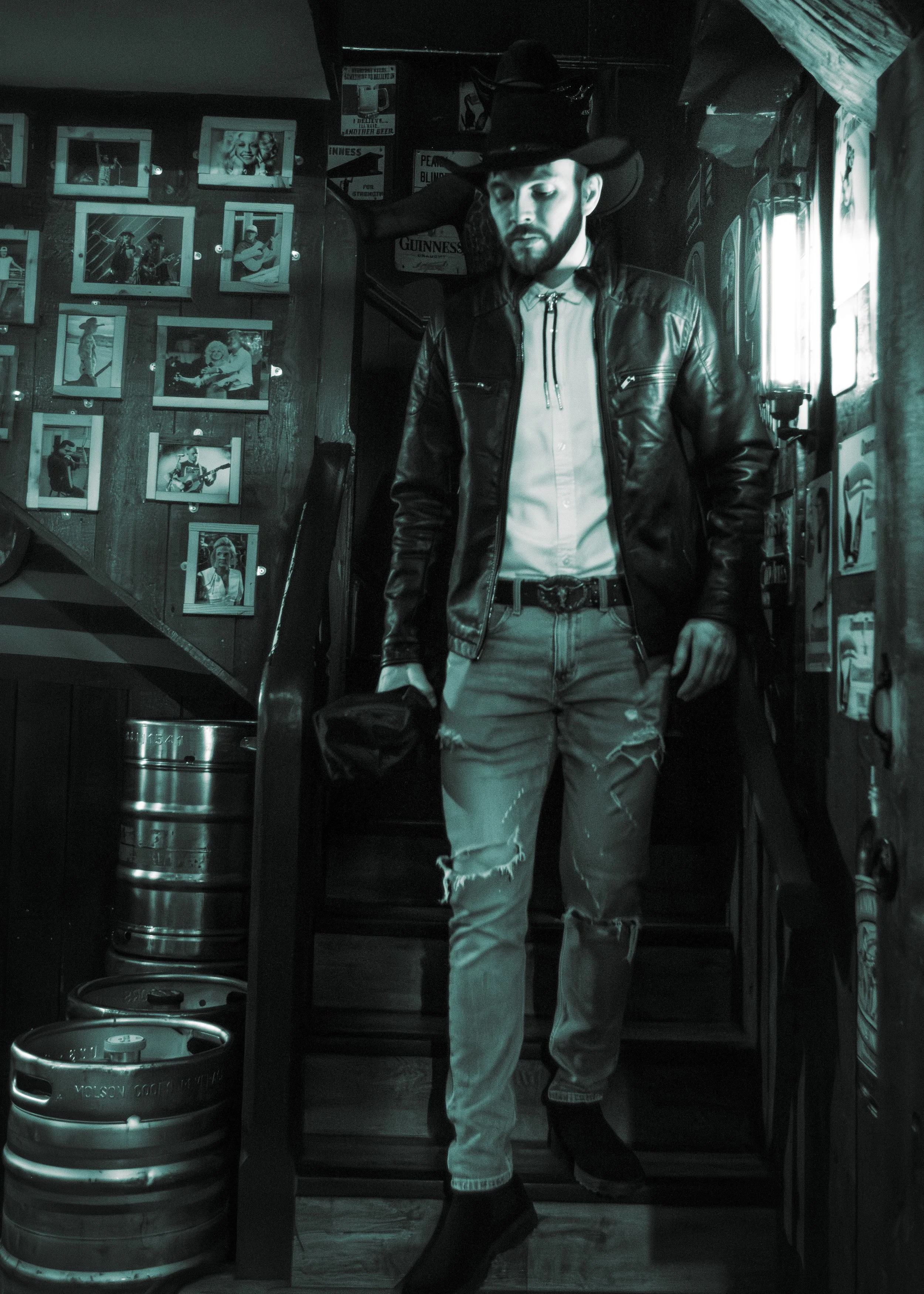 A man in a leather jacket, cowboy hat, and ripped jeans standing inside a dimly lit bar or pub with photos on the wall and beer kegs in the foreground.