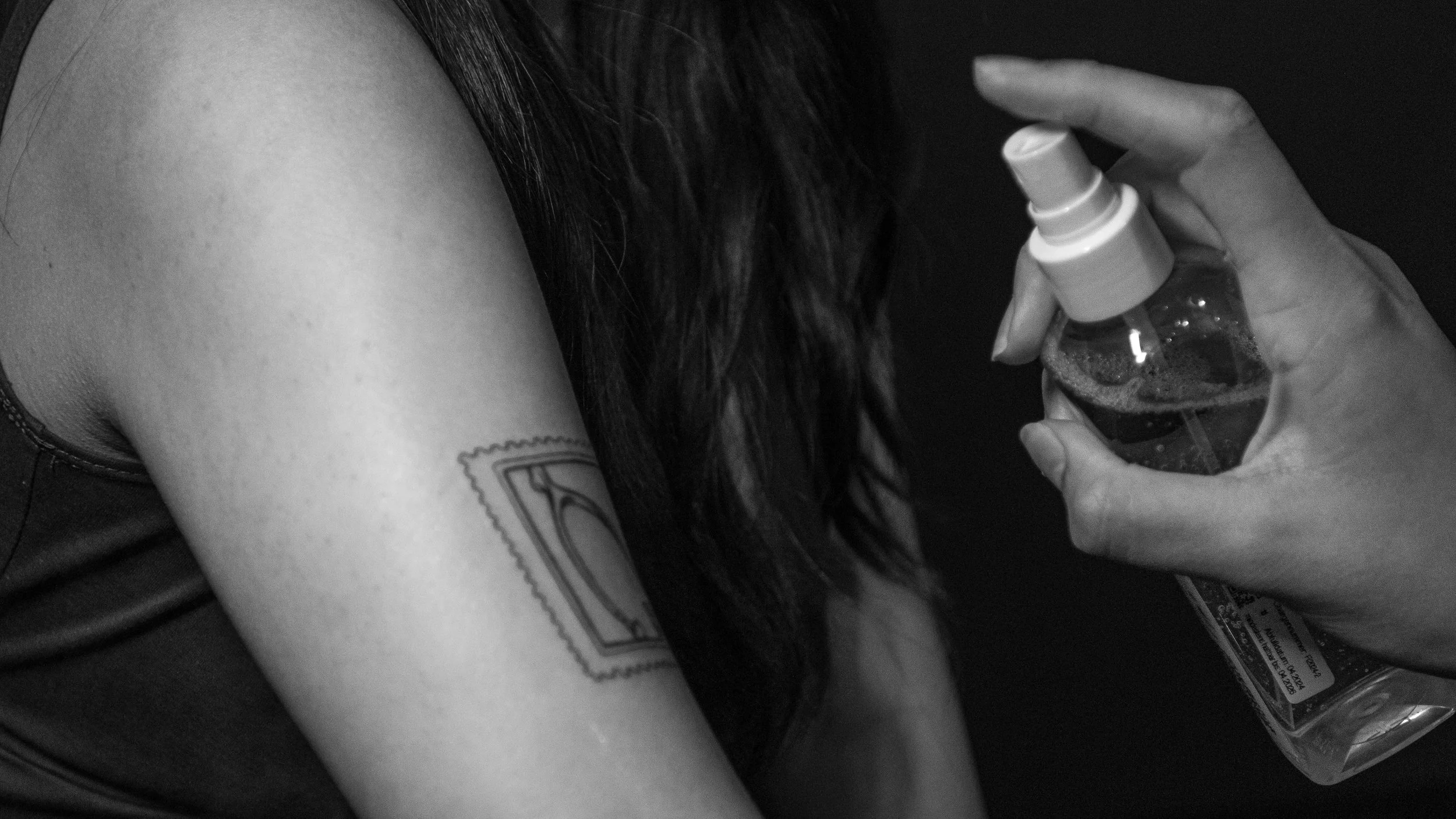 A person with long dark hair is holding a spray bottle near their shoulder, which has a tattoo of a rectangle with curved edges on their arm.