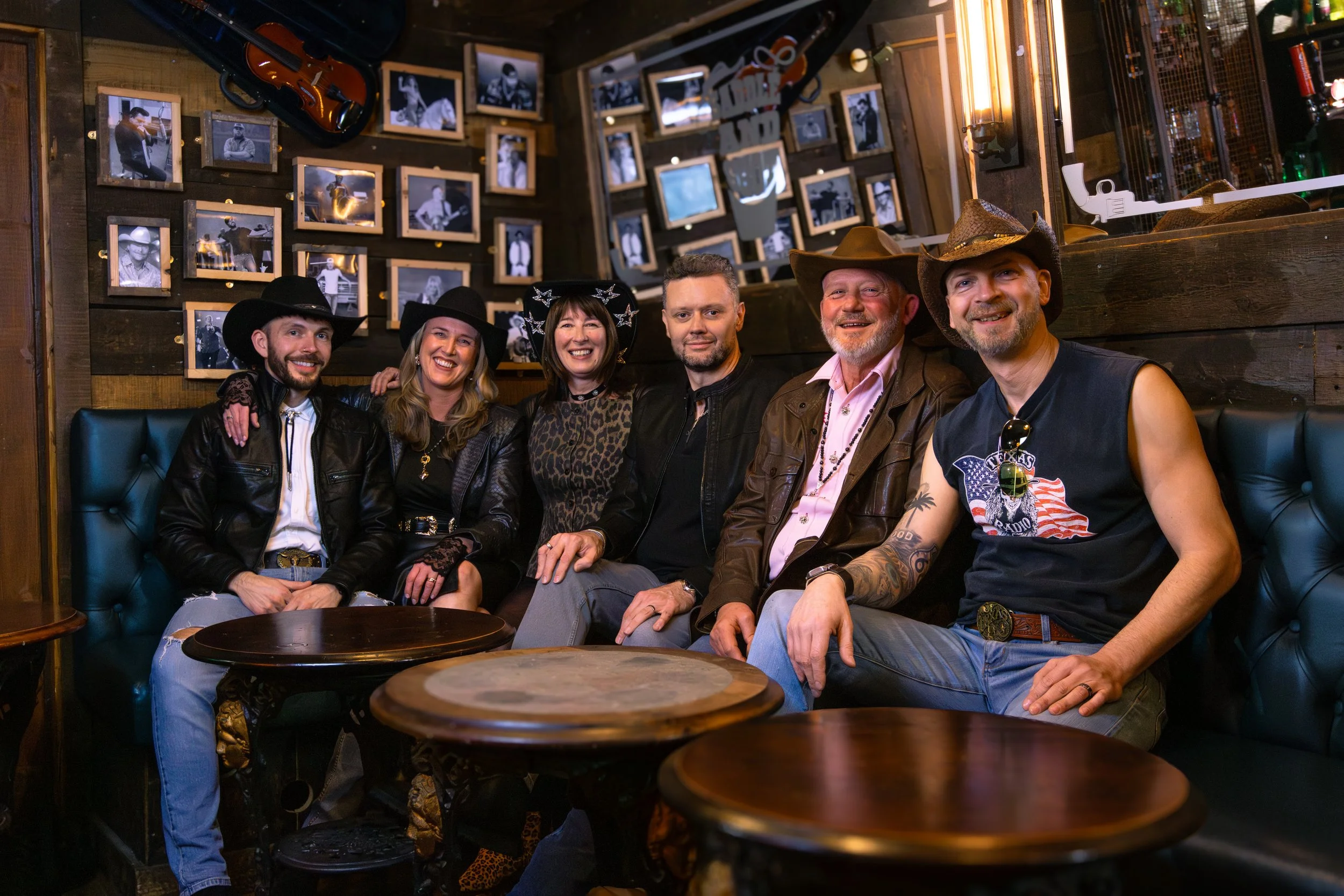 Six people sit on a leather sofa in a bar or restaurant with wooden walls decorated with framed black-and-white photos, guitars, and a skull mask. The group is smiling and dressed in casual and cowboy-themed attire.