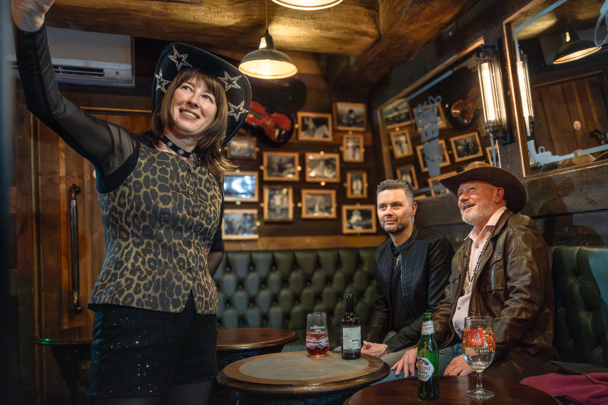 A woman wearing a black cowboy hat with star decorations taking a selfie in a bar or restaurant. Three men are sitting at a round table with drinks, smiling and looking at the woman. The background features photo frames on the wall and warm lighting.