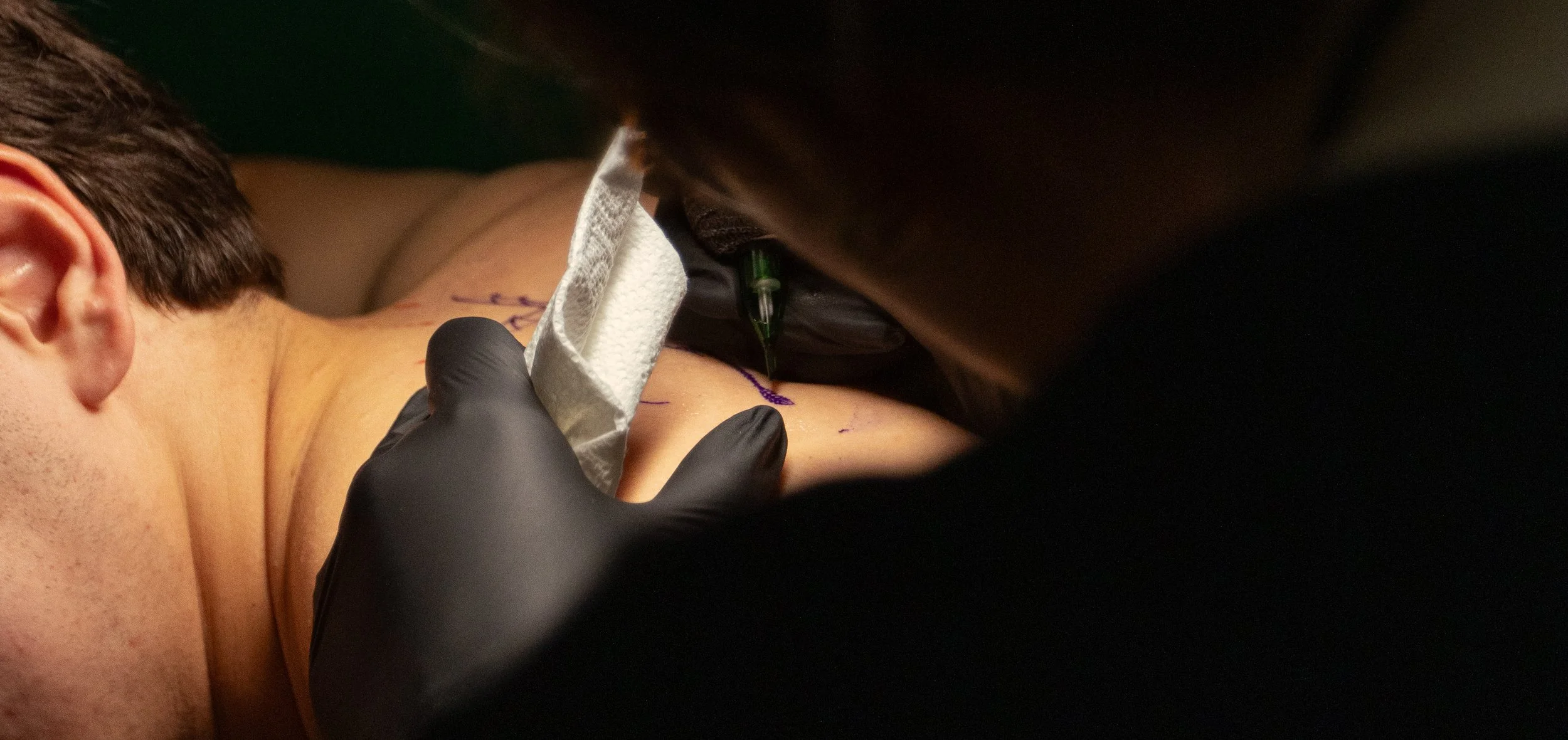 A person receiving a tattoo on their upper back or shoulder area, with a tattoo needle and ink visible, and the tattoo artist wearing black gloves.