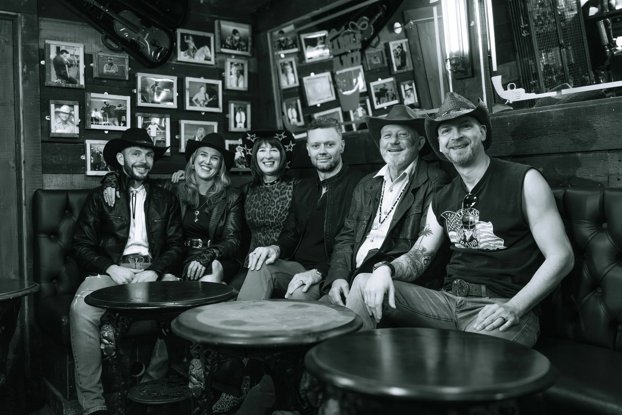 Group of six people sitting together in a bar or restaurant, smiling and wearing cowboy hats, with framed photos on the wall behind them.