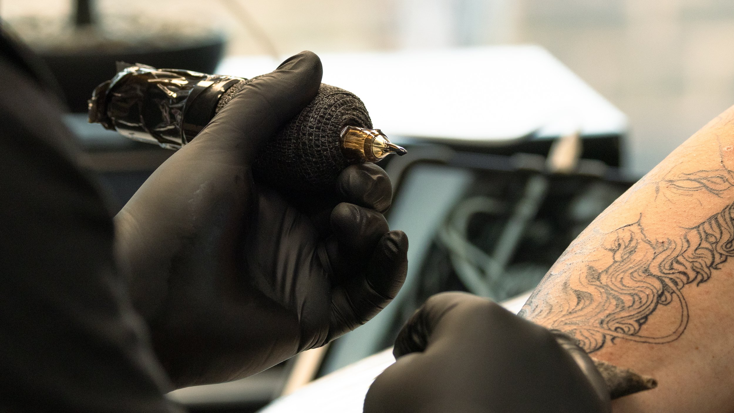 A tattoo artist wearing black gloves is holding a tattoo machine, tattooing a design on a person's arm. The person's forearm is visible with a detailed tattoo line work.