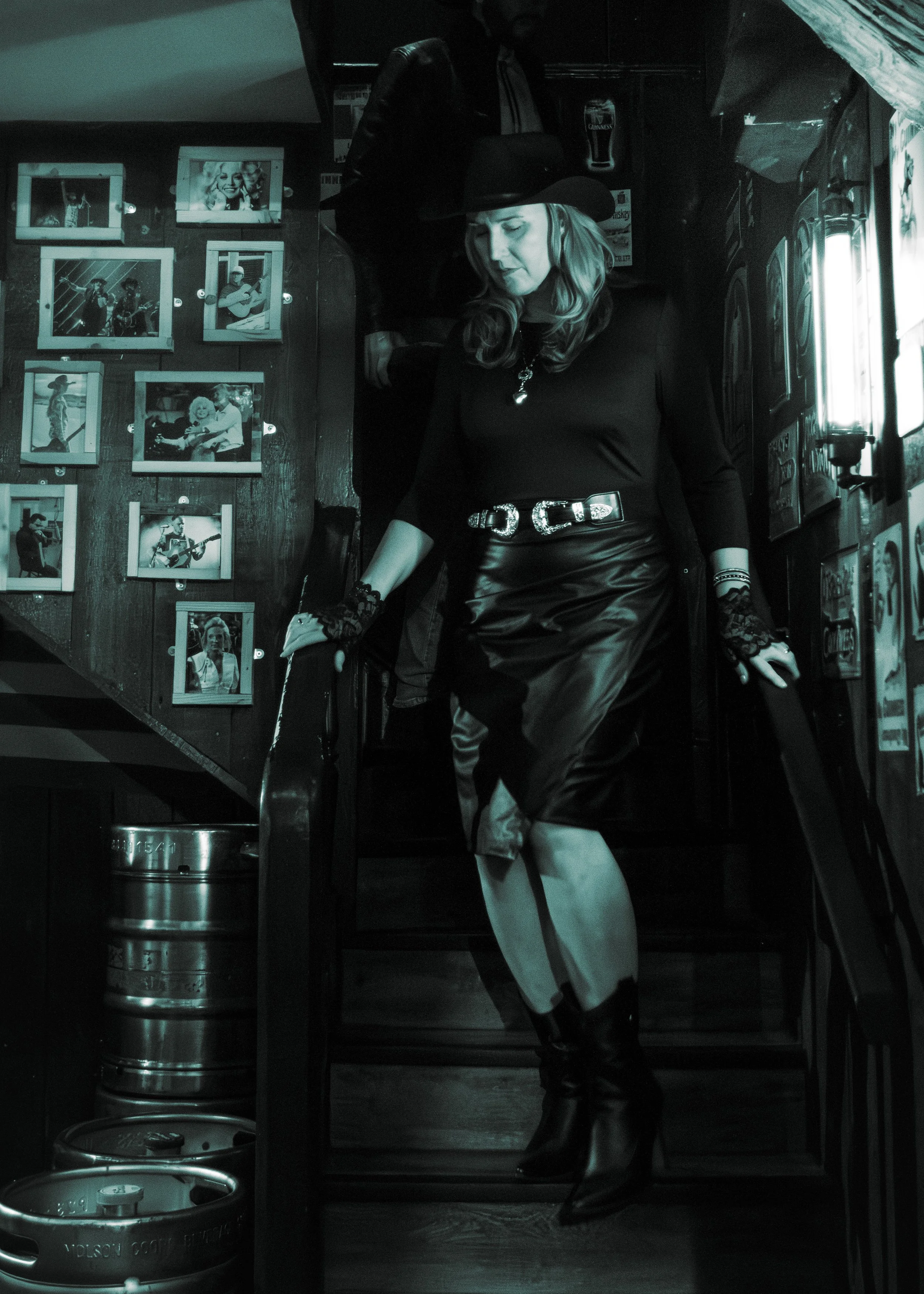A woman dressed as a witch, wearing a black hat and black leather skirt, standing inside a bar or pub near a wall decorated with black-and-white photos of musicians. She is looking down, with one hand resting on a railing and the other near her waist