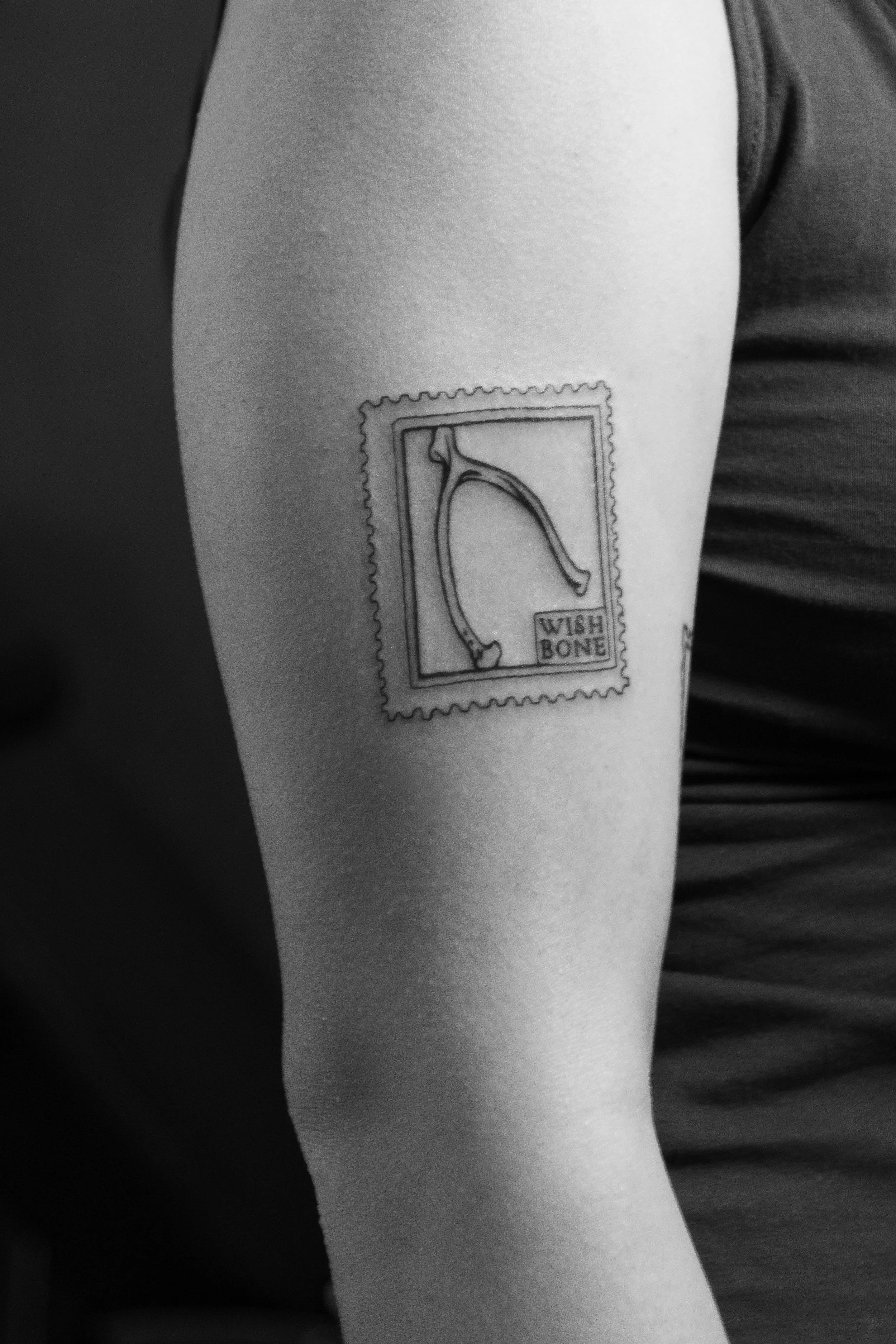 A person's upper arm with a tattoo of a postage stamp featuring a high-heeled shoe and the words 'Wish Bone'.