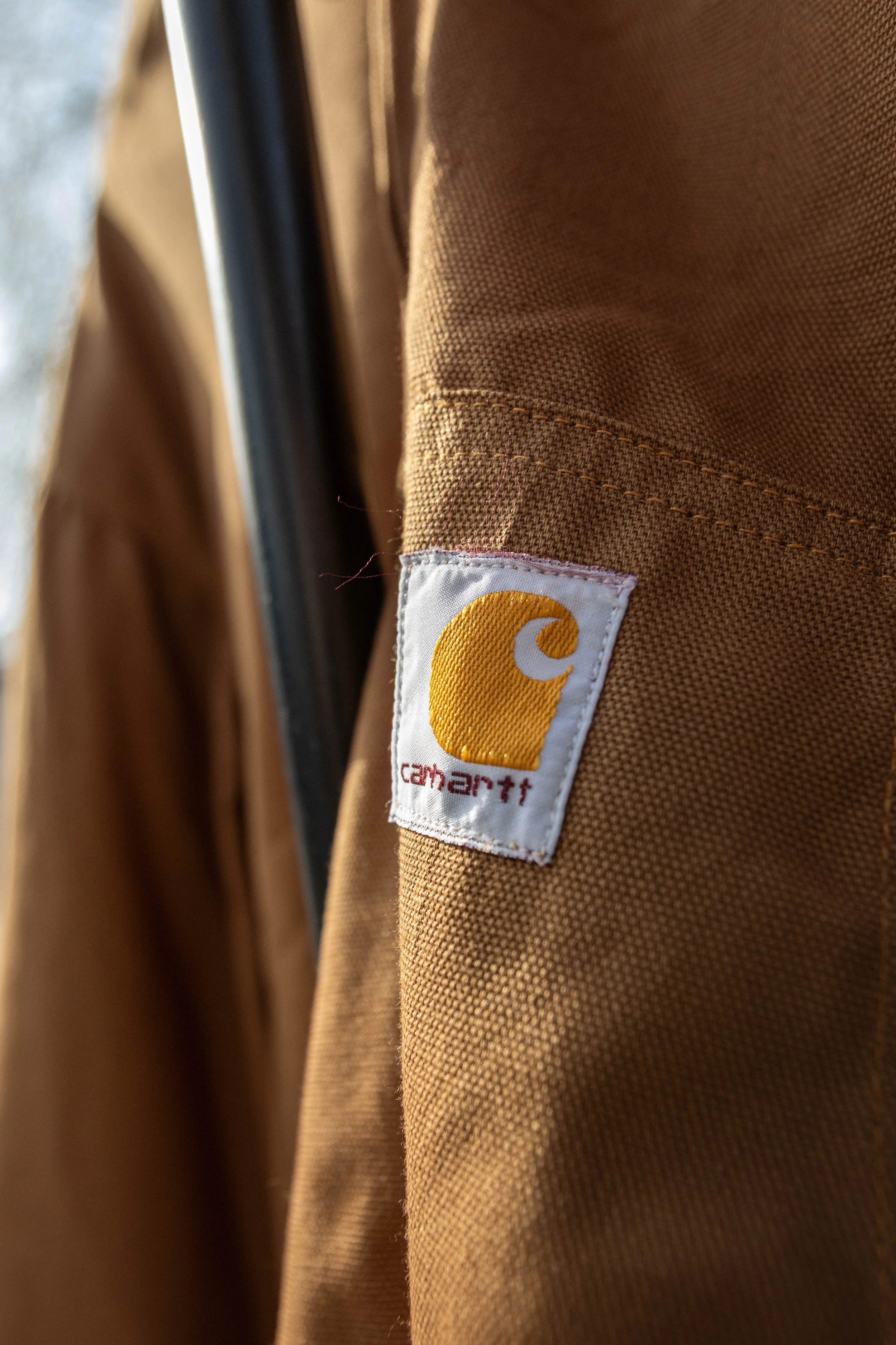 Close-up of a brown Carhartt work jacket showing a white label with the Carhartt logo and name, with the fabric textured and detailed.