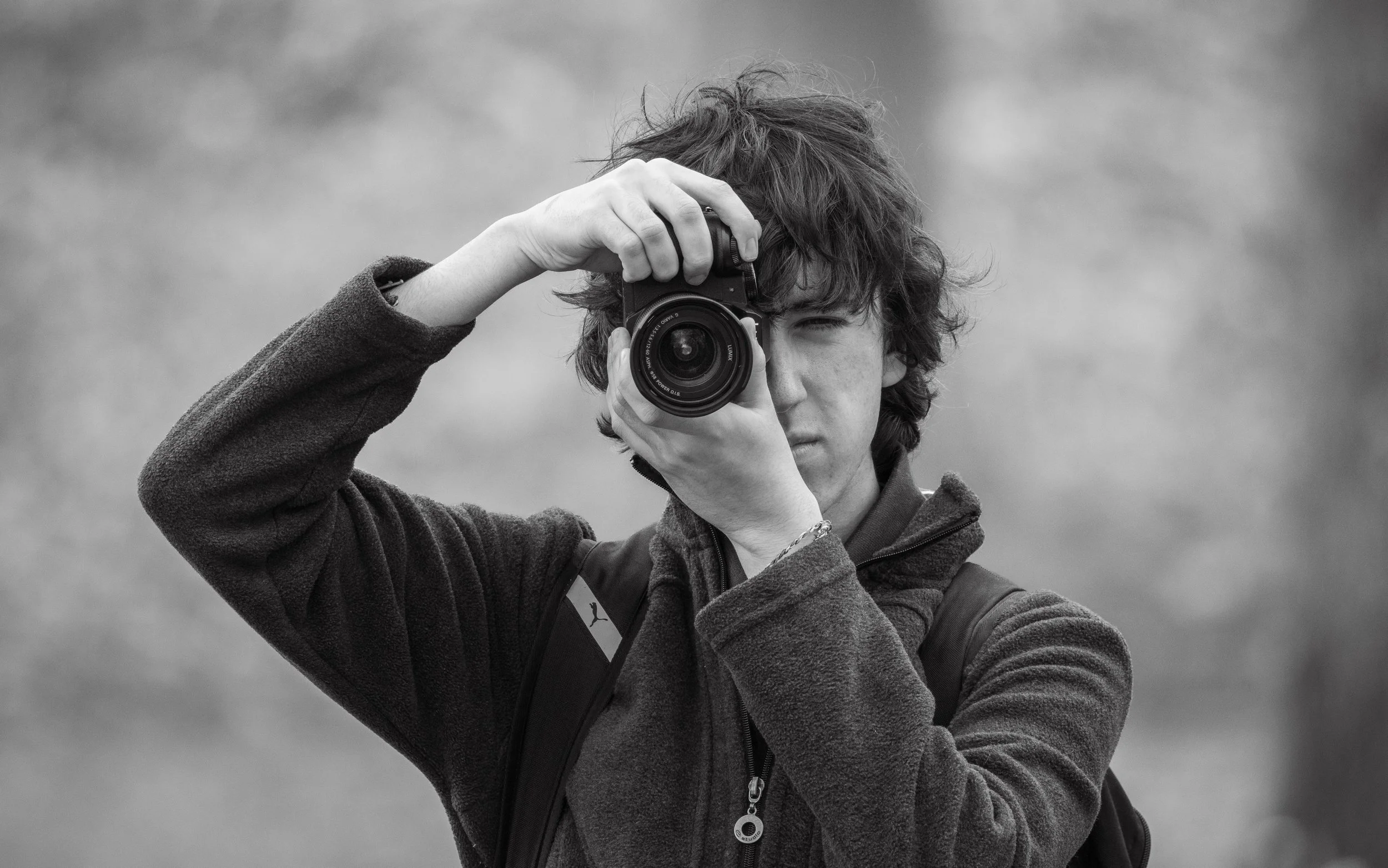 Young person with curly hair taking a photo with a camera outdoors, wearing a jacket and a backpack.
