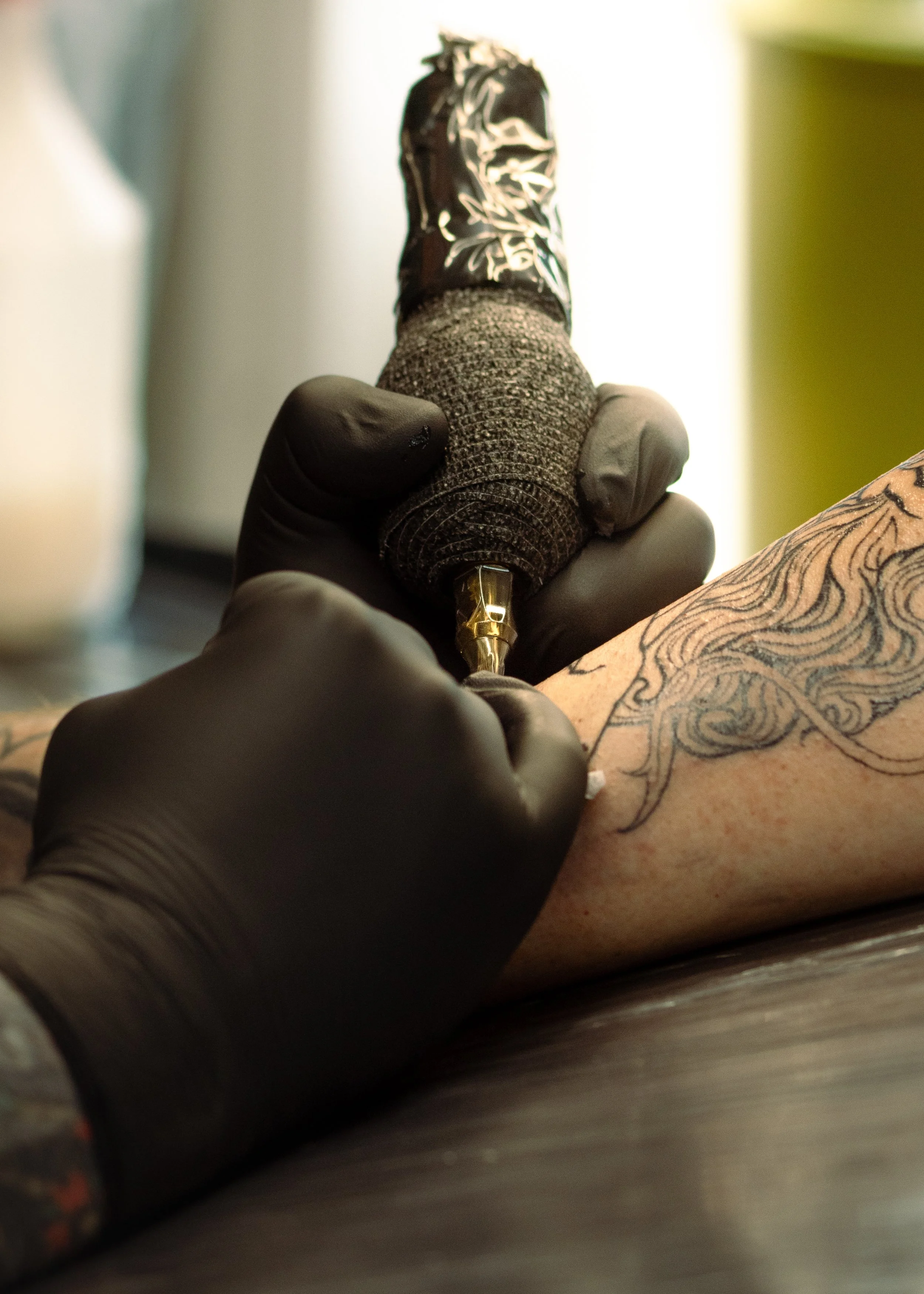 A tattoo artist tattoos a design on a person's arm using a tattoo gun, with the person lying down on a table.
